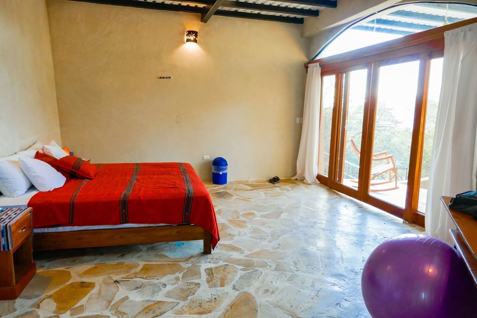 Un dormitorio con cama, una mesita, una silla y un amplio ventanal.