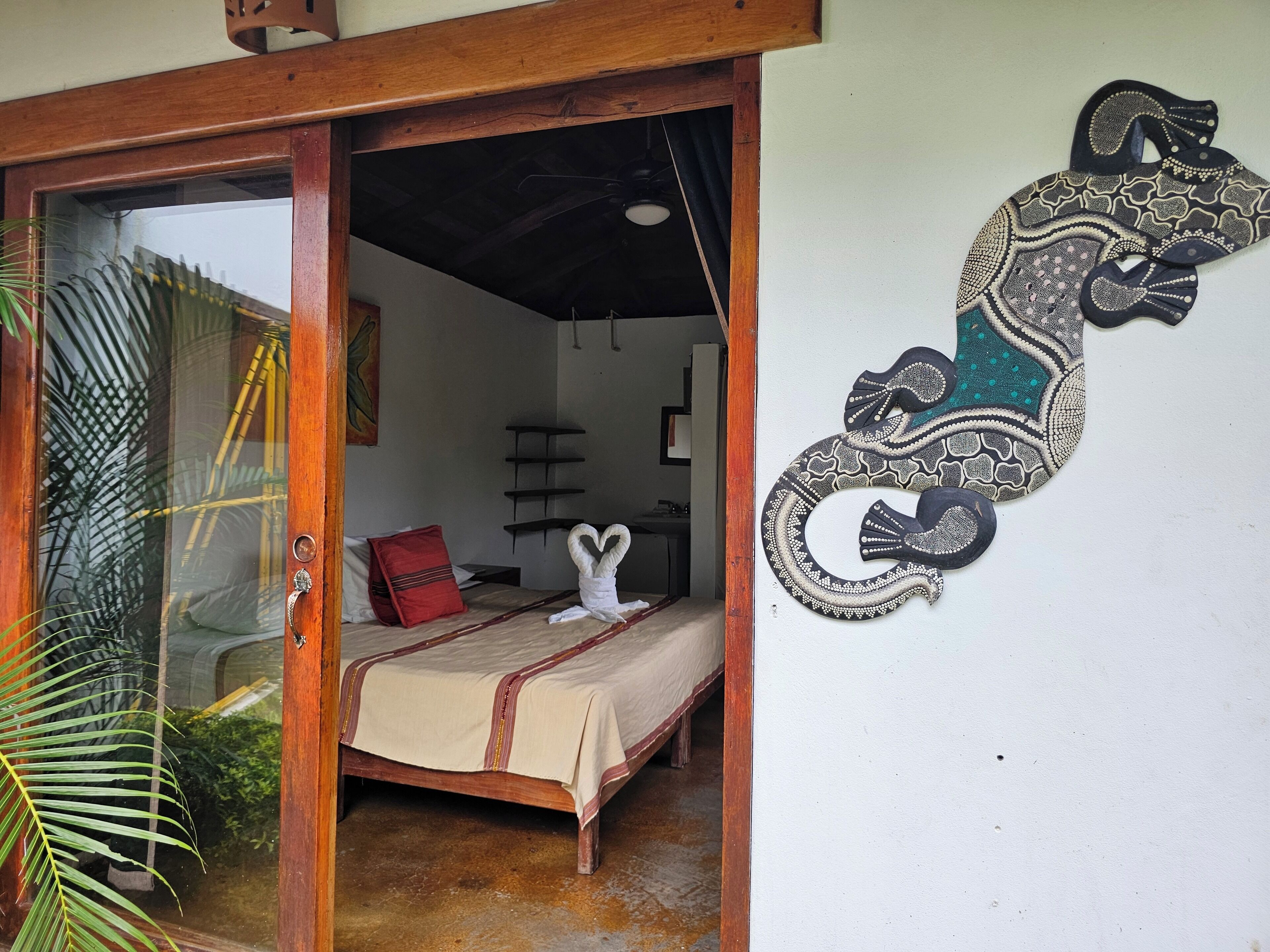 A room with a bed, a wooden door, a wall-mounted decorative piece, and a view of the outside.