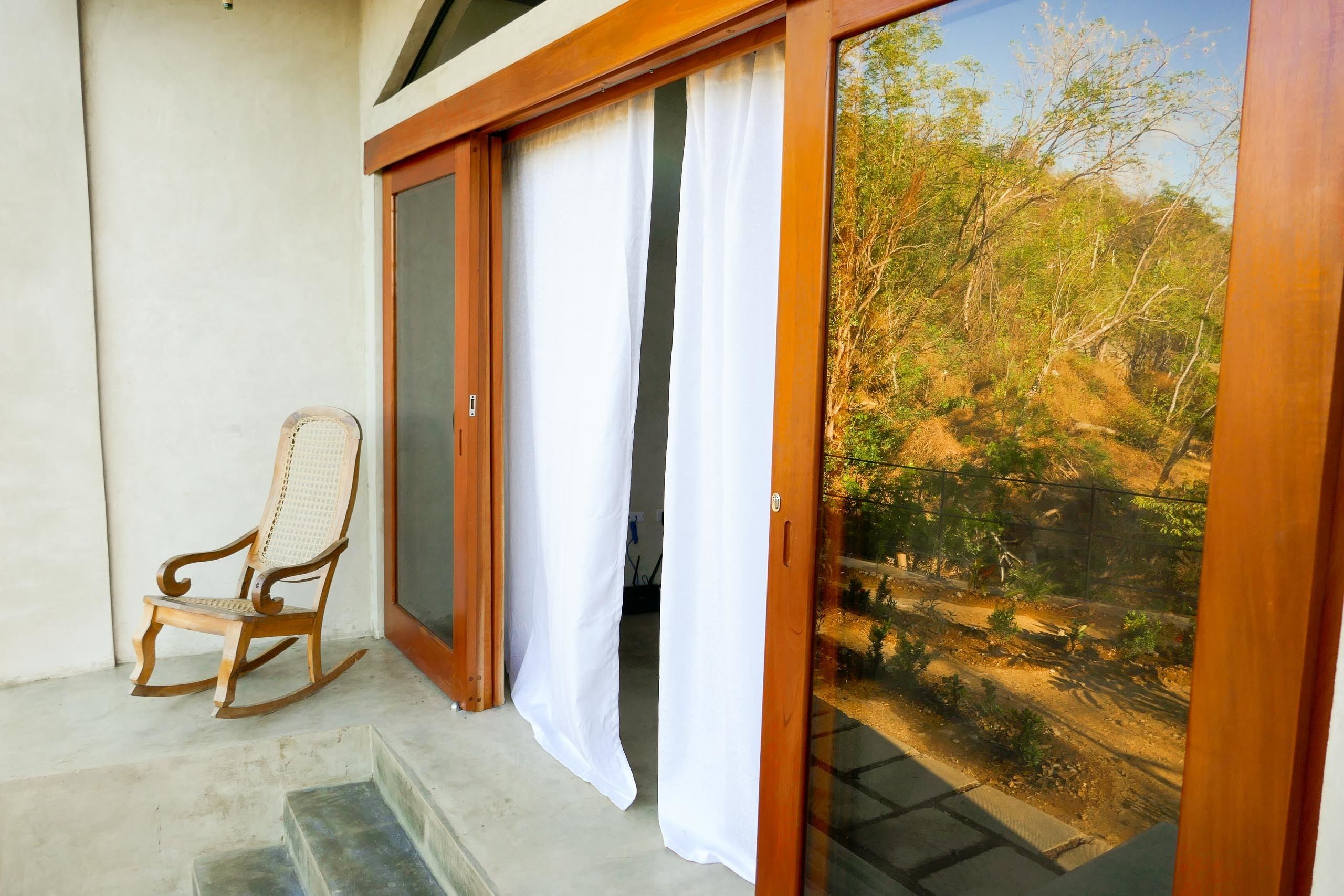 A wooden rocking chair outside a sliding glass door leading to a garden with a dirt path and trees.