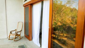 A wooden rocking chair outside a sliding glass door leading to a garden with a dirt path and trees.