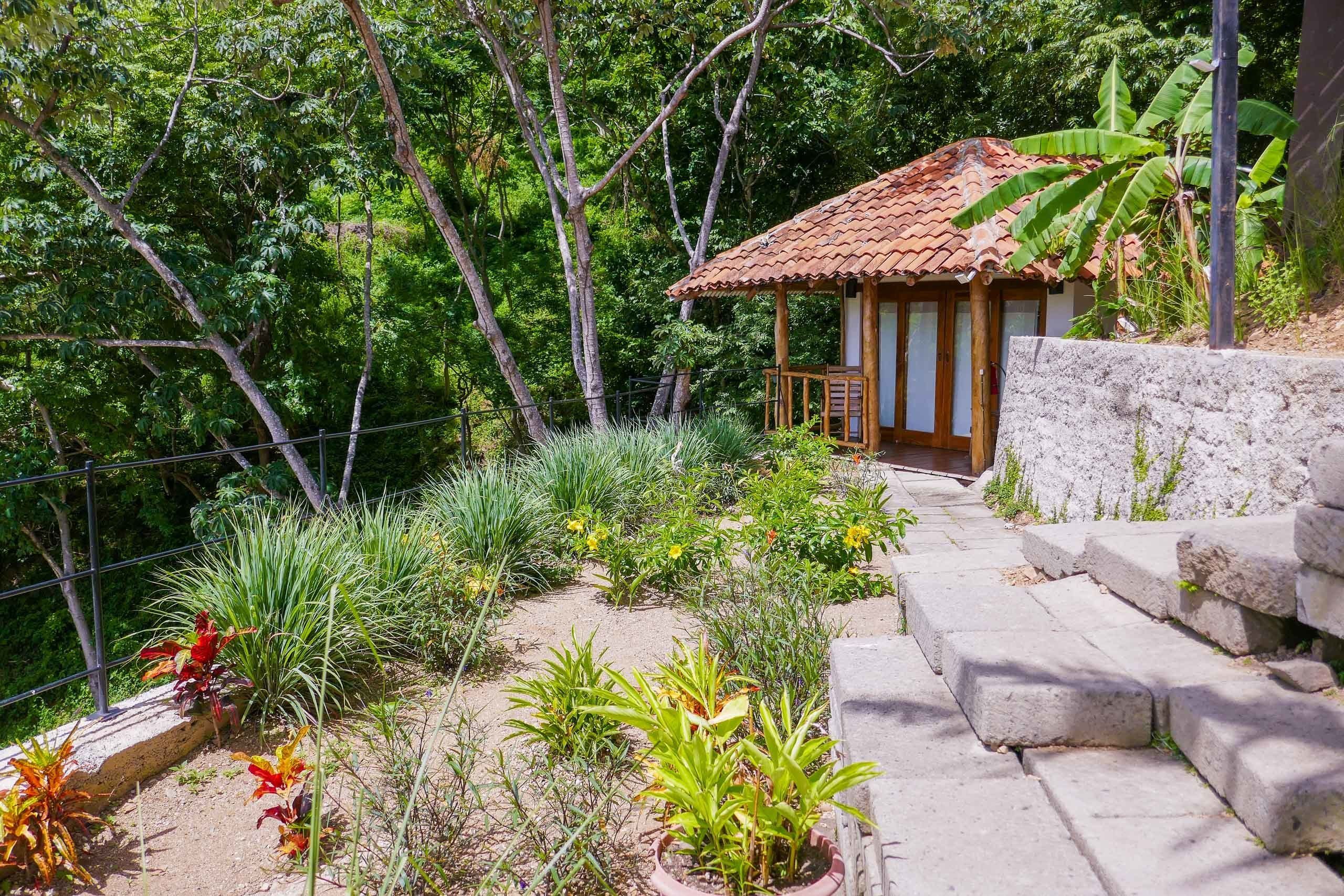 A small wooden structure with a tiled roof, surrounded by a garden with various plants and a stone wall.