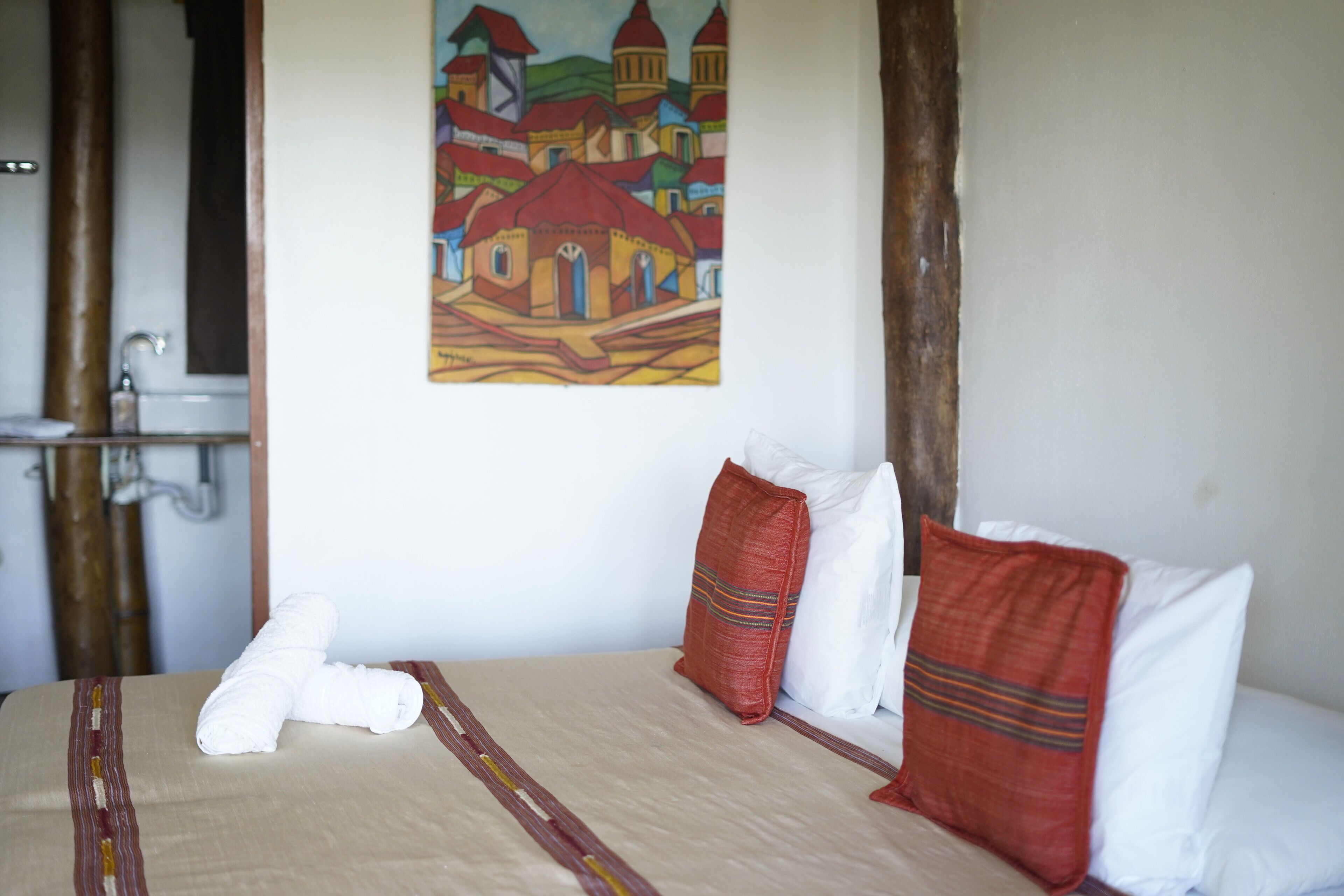 A hotel room with a bed, pillows, a painting on the wall, and a bathroom with a sink.
