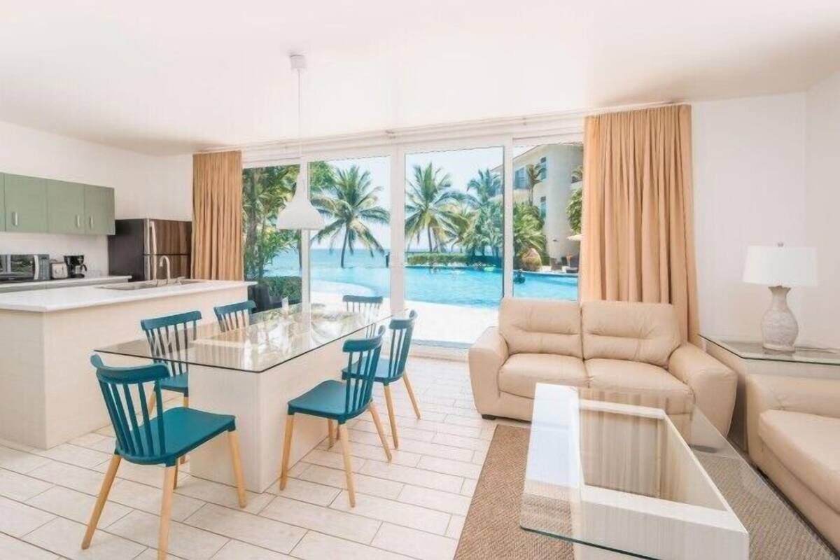 Un soggiorno moderno con un tavolino in vetro, un divano beige e un'area pranzo con un tavolo in vetro e sedie blu. Si intravede una piscina e palme attraverso ampie finestre.