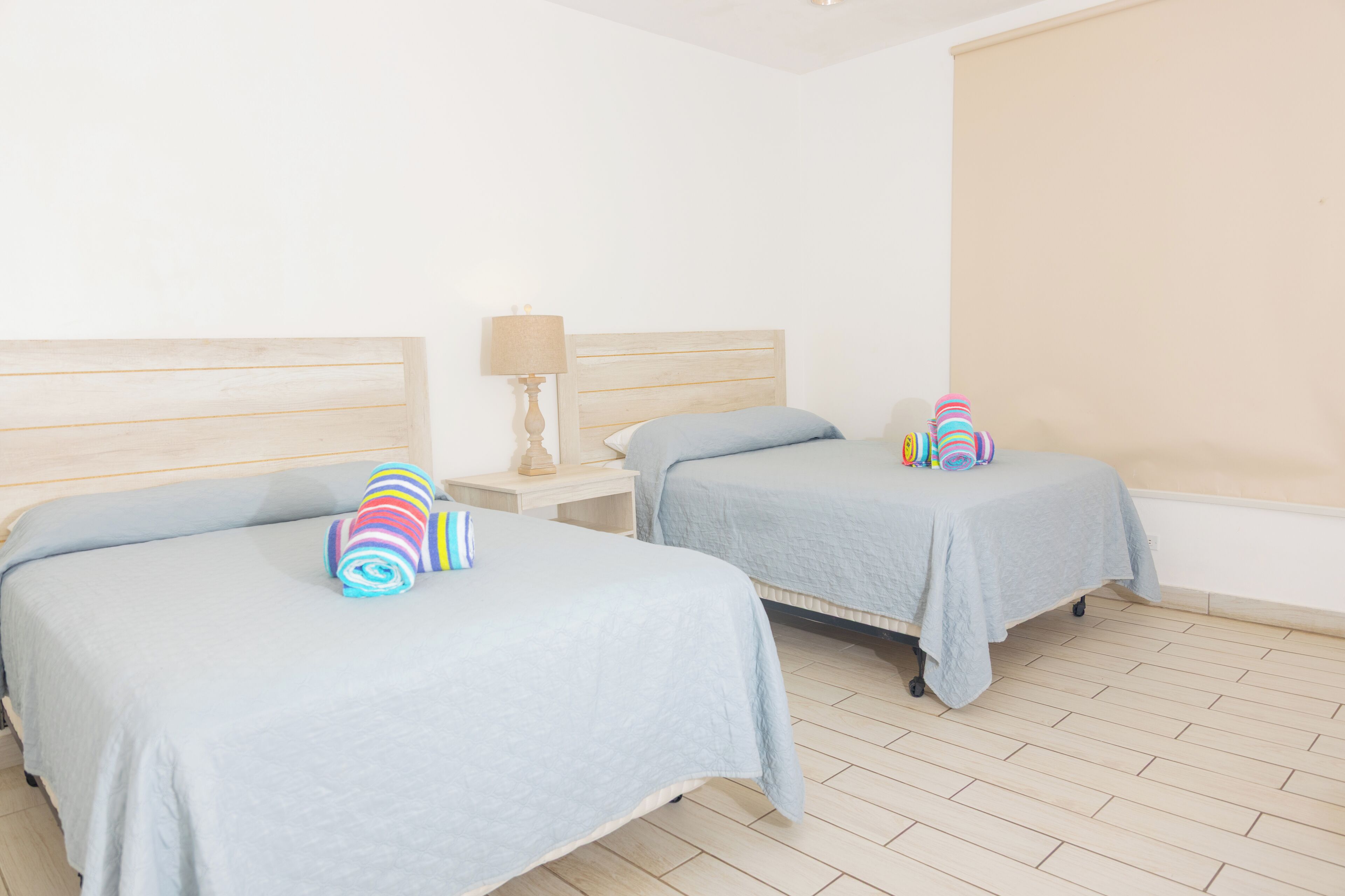 A hotel room with two beds, each with a blue bedspread and striped pillows. There is a nightstand with a lamp between the beds.