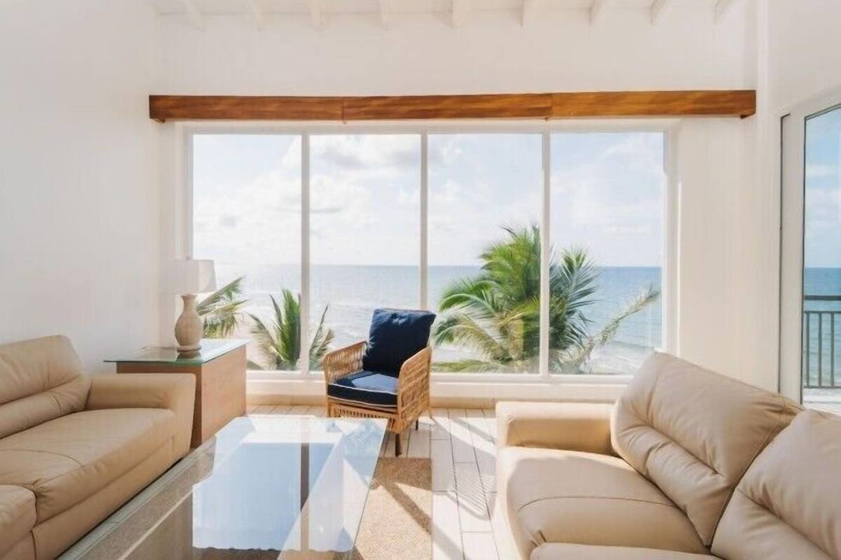 Un ampio soggiorno con una grande finestra che offre vista sull'oceano e sulle palme. Sono presenti due divani beige, un tavolino in vetro e una poltrona in vimini con un cuscino blu.