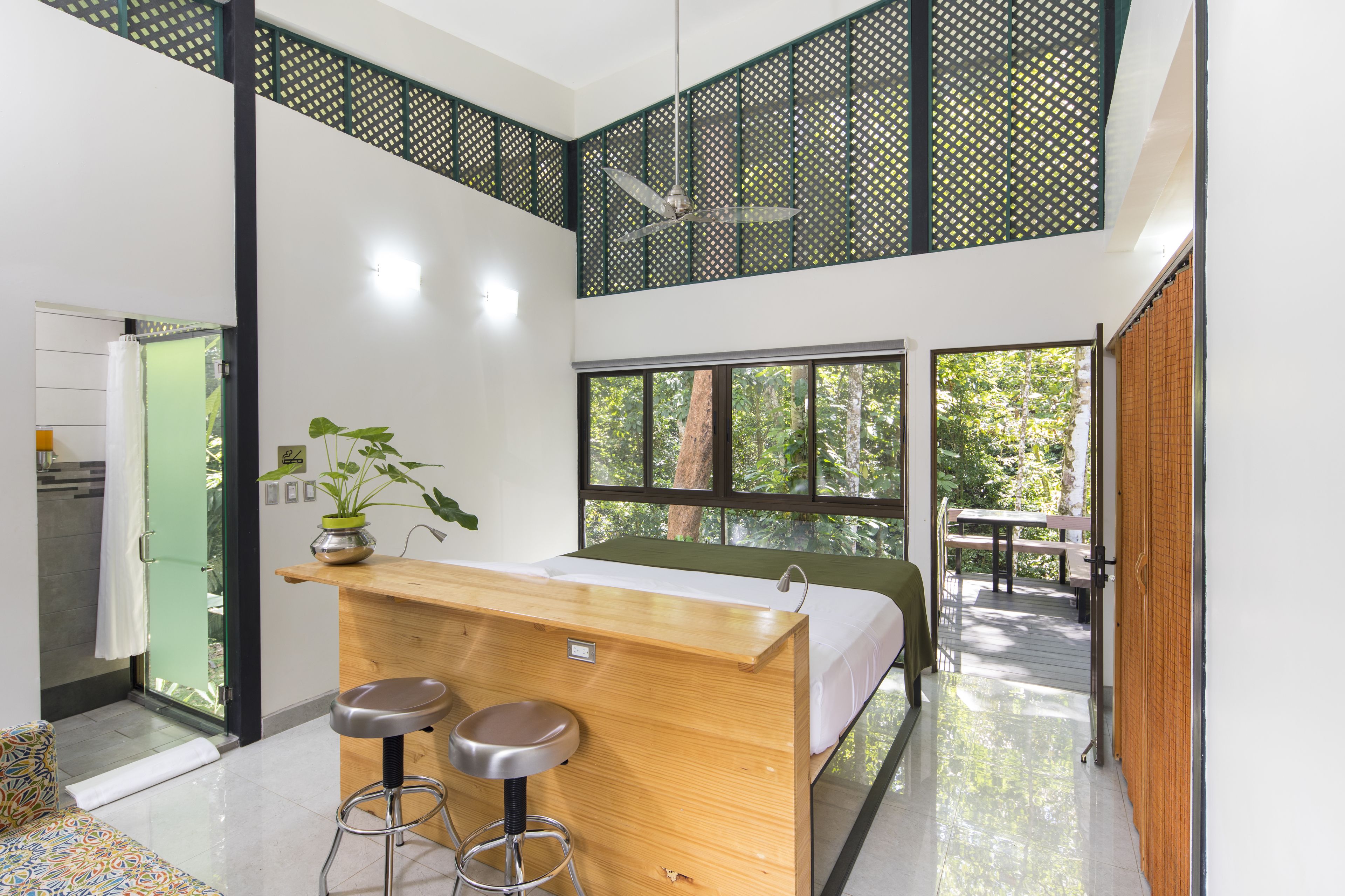 A modern room with a wooden desk, two bar stools, a bed, and large windows offering a view of trees.