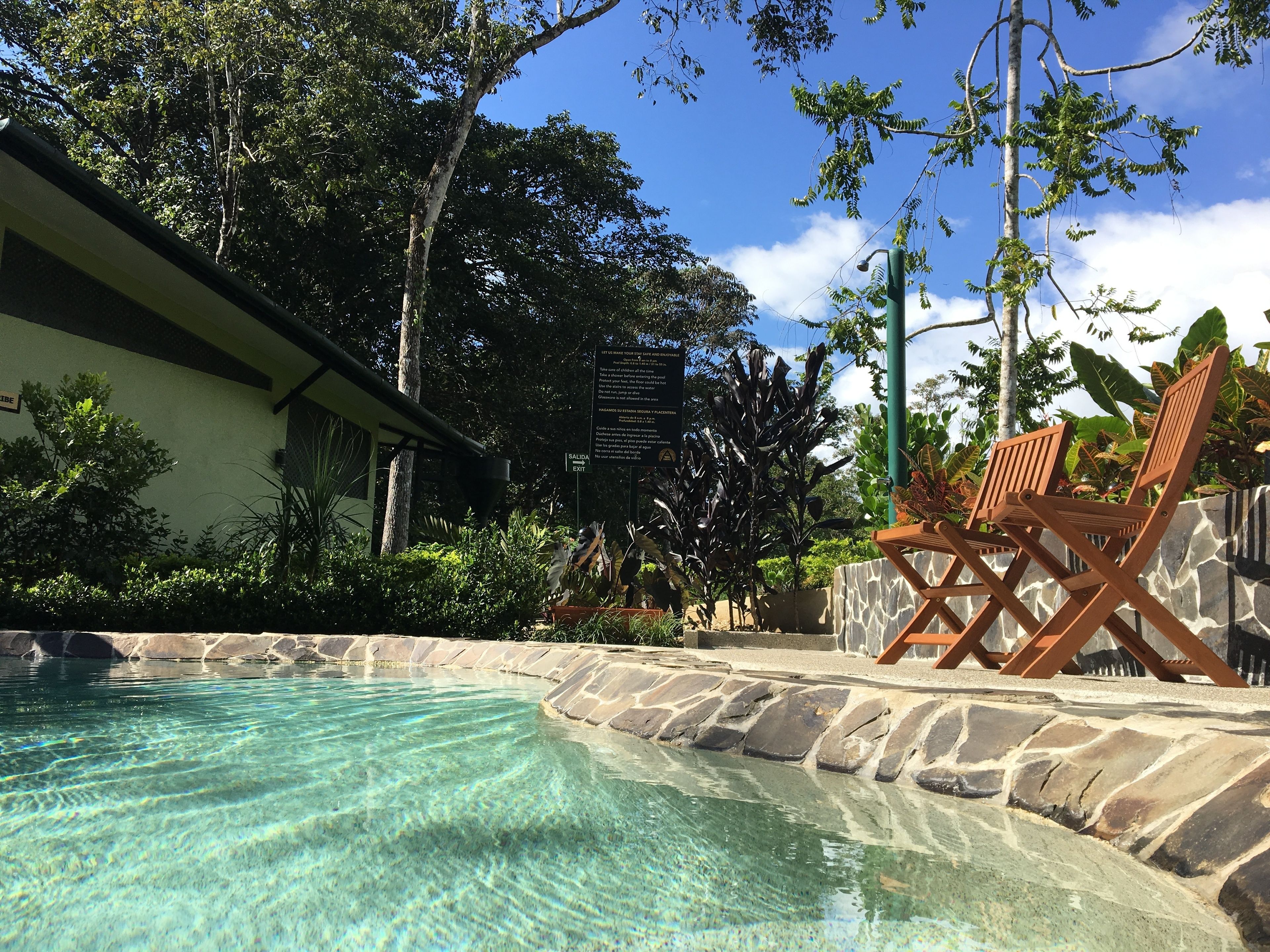 Une piscine aux eaux cristallines, bordée de pierres, entourée d’une végétation luxuriante et agrémentée d’une terrasse en bois avec des chaises.