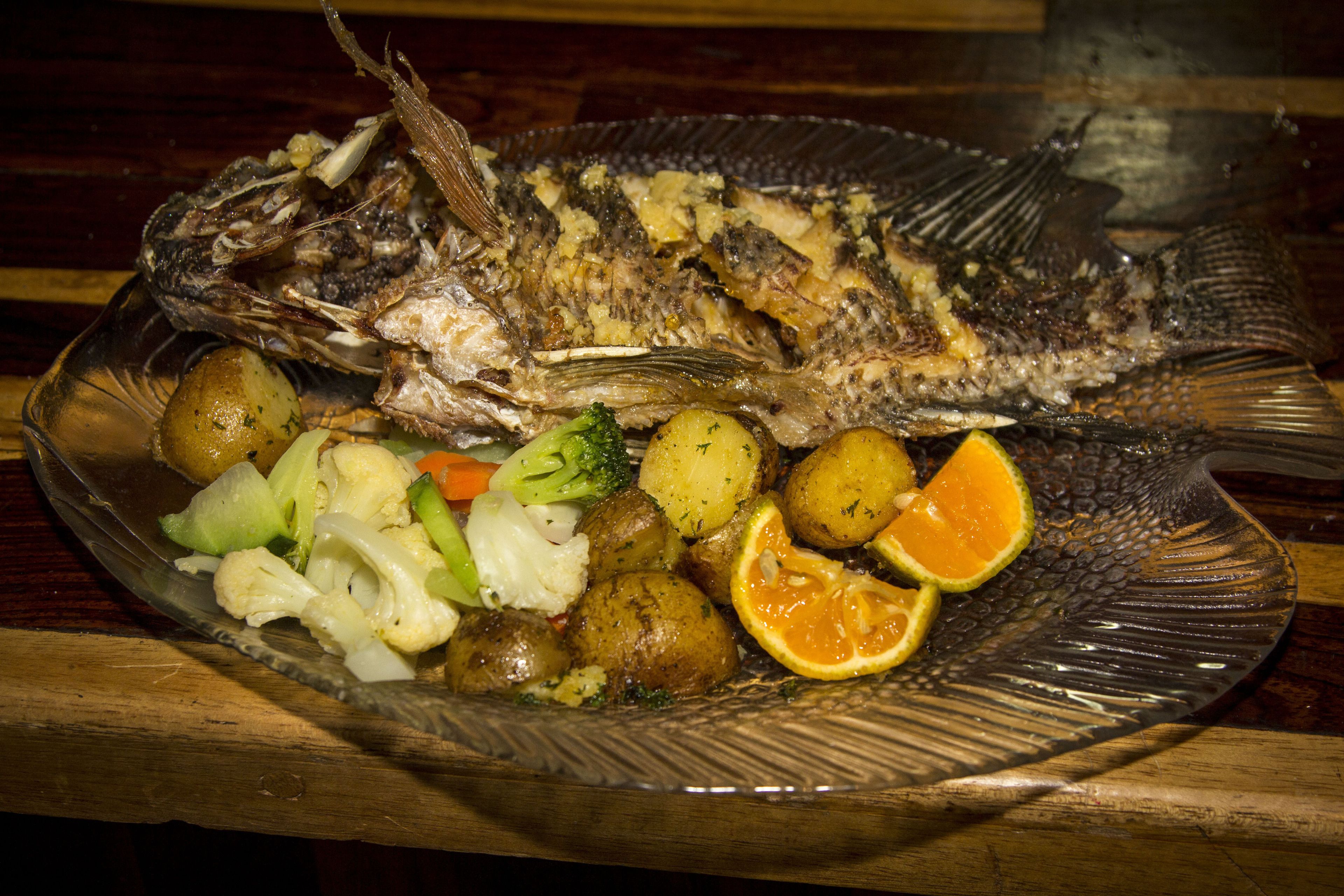 Un pescado entero en un plato con patatas, coliflor y una rodaja de naranja.