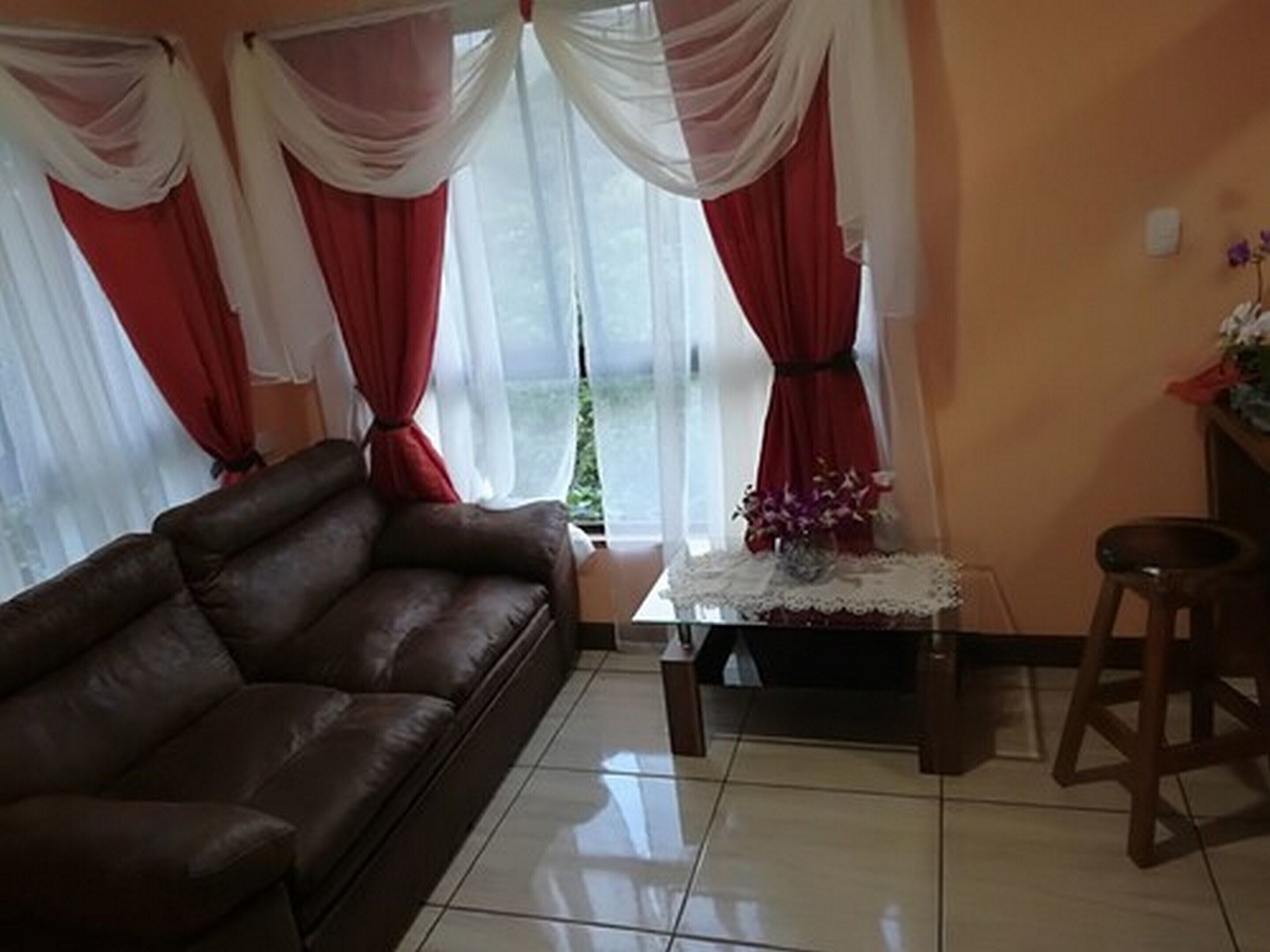 A living room with a brown leather sofa, a coffee table with a vase of flowers, and red and white curtains.