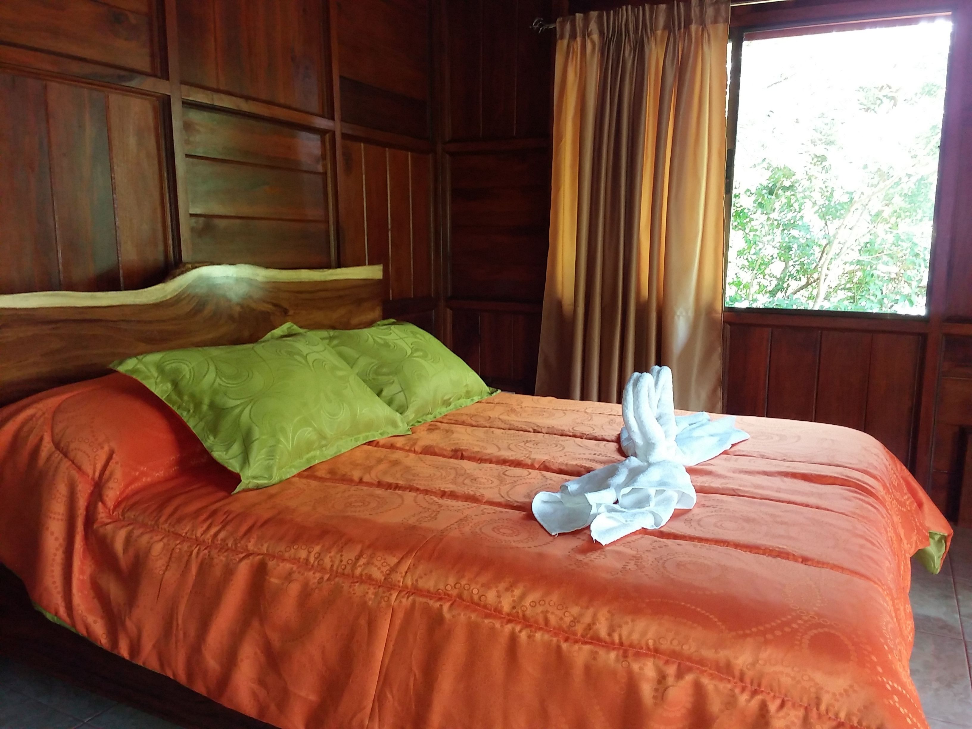 A wooden room with a bed, green and orange bedding, and a window with curtains.