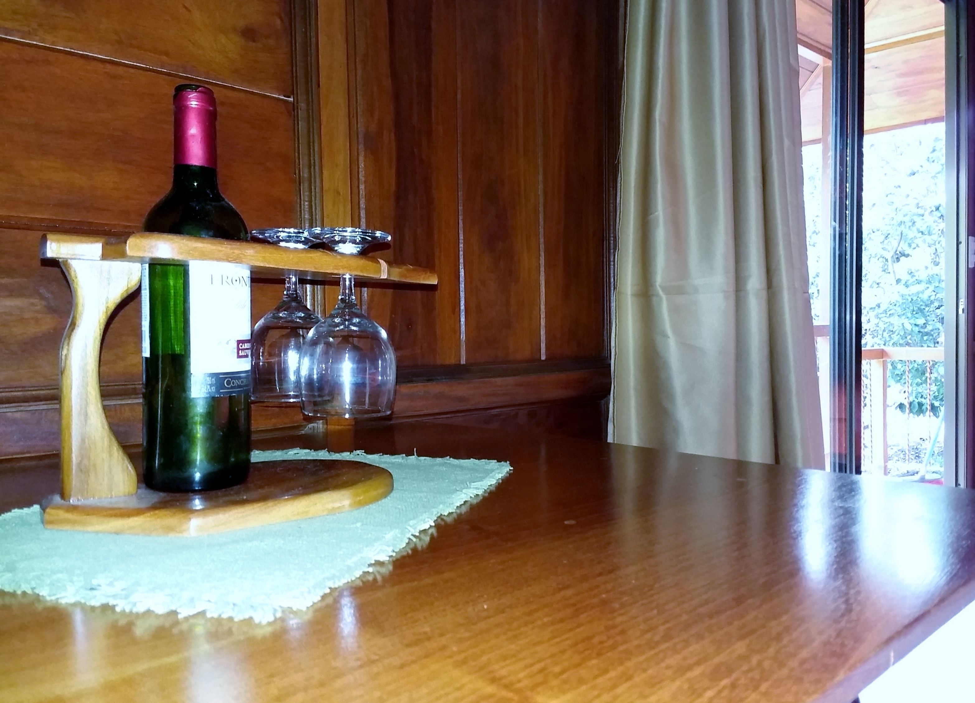 Uma adega de madeira com uma garrafa e copos sobre uma mesa perto de uma janela com cortinas.