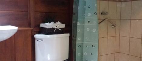 A bathroom with a toilet, a sink, a trash bin, a shower curtain, and a shelf with a plant.