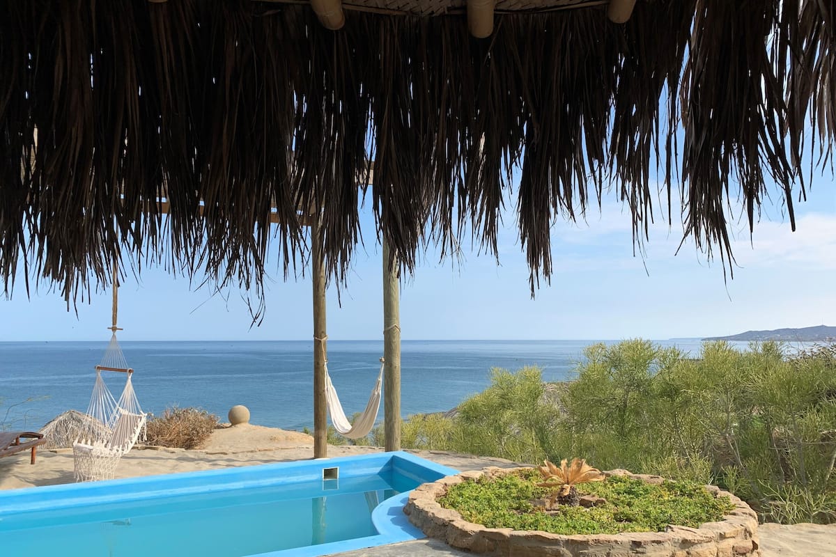 A pool area with a view of the sea, a hammock, and a thatched roof structure.