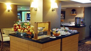 Zona de bufé con diversas frutas y bebidas, máquina de café y un área de descanso con mesa y sillas.