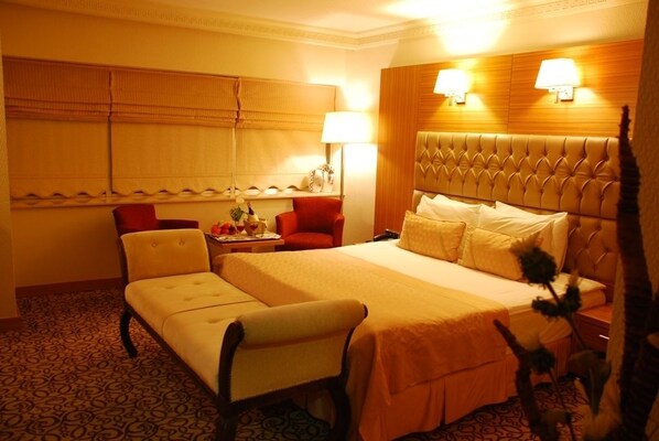 A hotel room with a large bed, a sitting area, a desk, and a window with blinds. - Emir Royal Hotel (Adana)