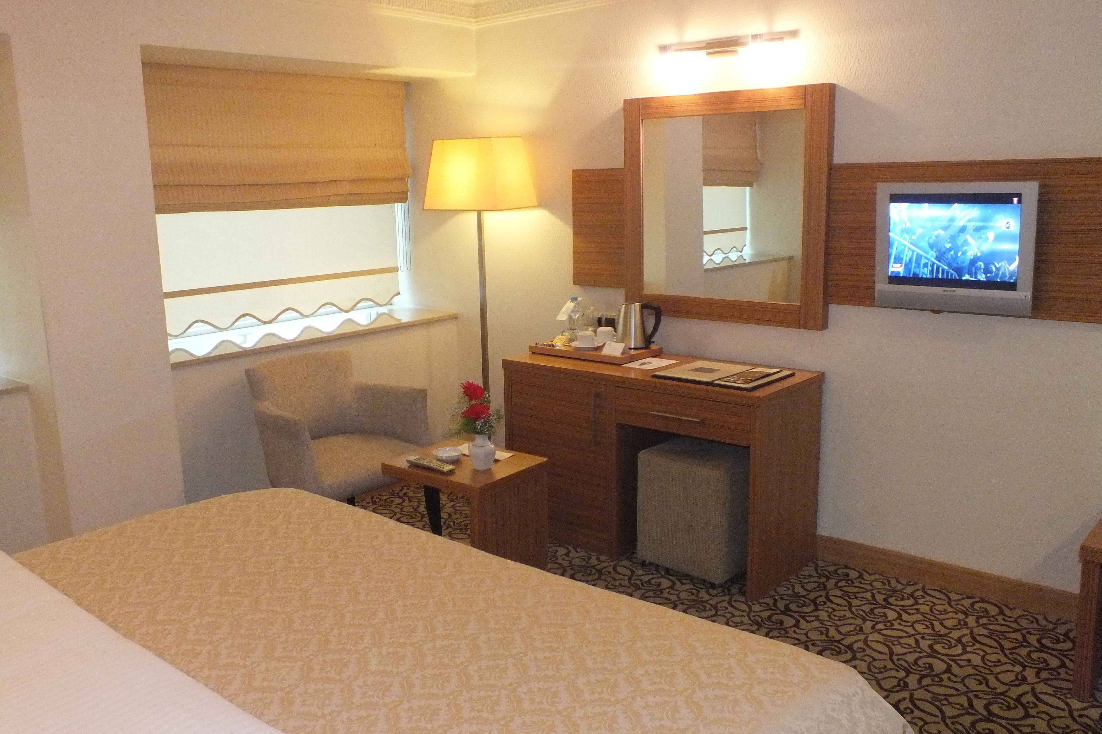 A hotel room with a bed, a desk with a lamp, a television, and a window with blinds.
