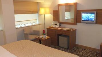 A hotel room with a bed, a desk with a lamp, a television, and a window with blinds.