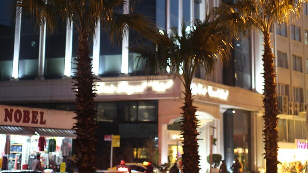 Un hotel con fachada moderna, palmeras en primer plano y un escaparate visible con la palabra 'NOBEL' en la planta baja.
