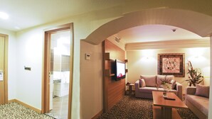 Habitación de hotel con sala de estar, sofá, mesa de centro, televisor montado en la pared y puerta de madera.