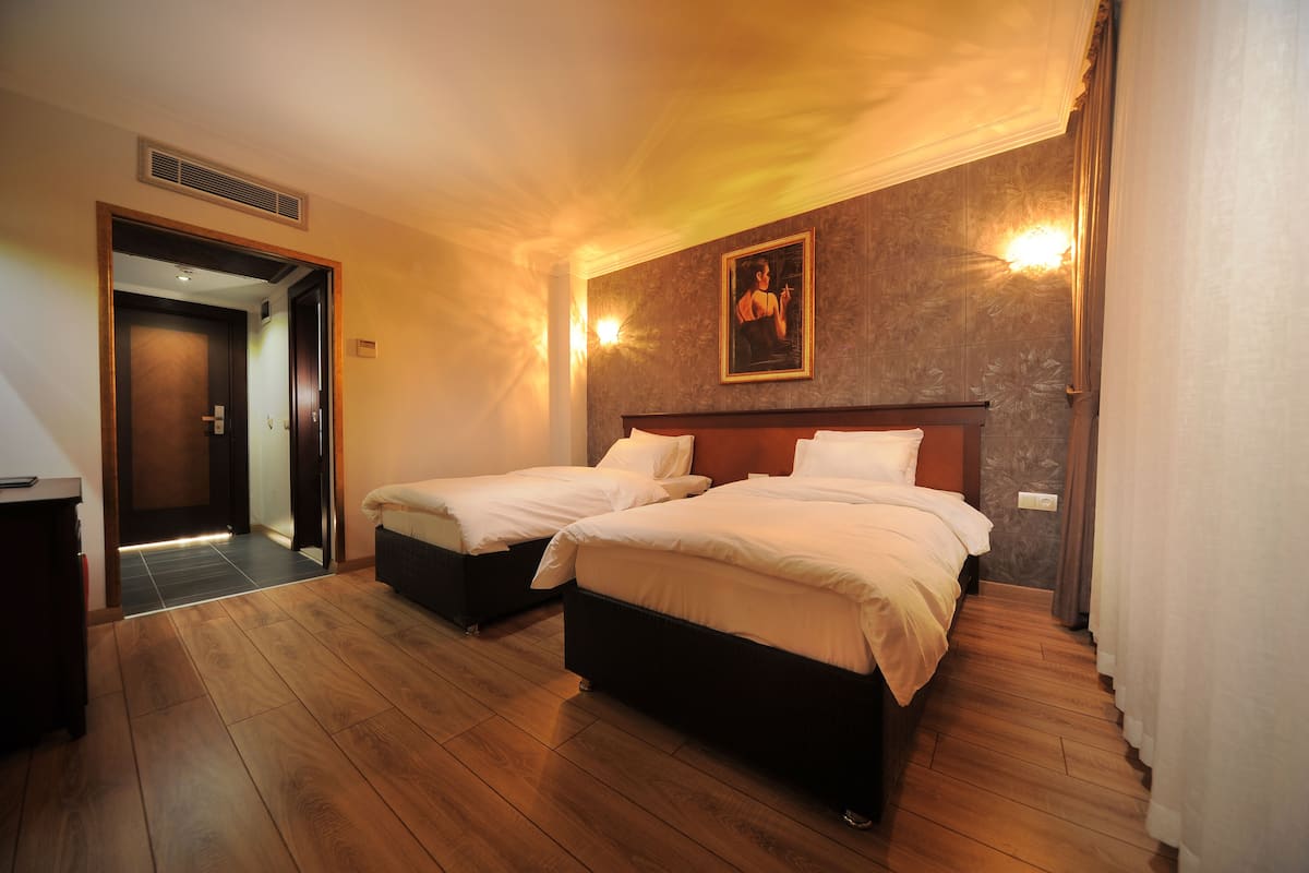 A hotel room with two beds, wooden flooring, a door leading to another room, and a wall-mounted light fixture.
