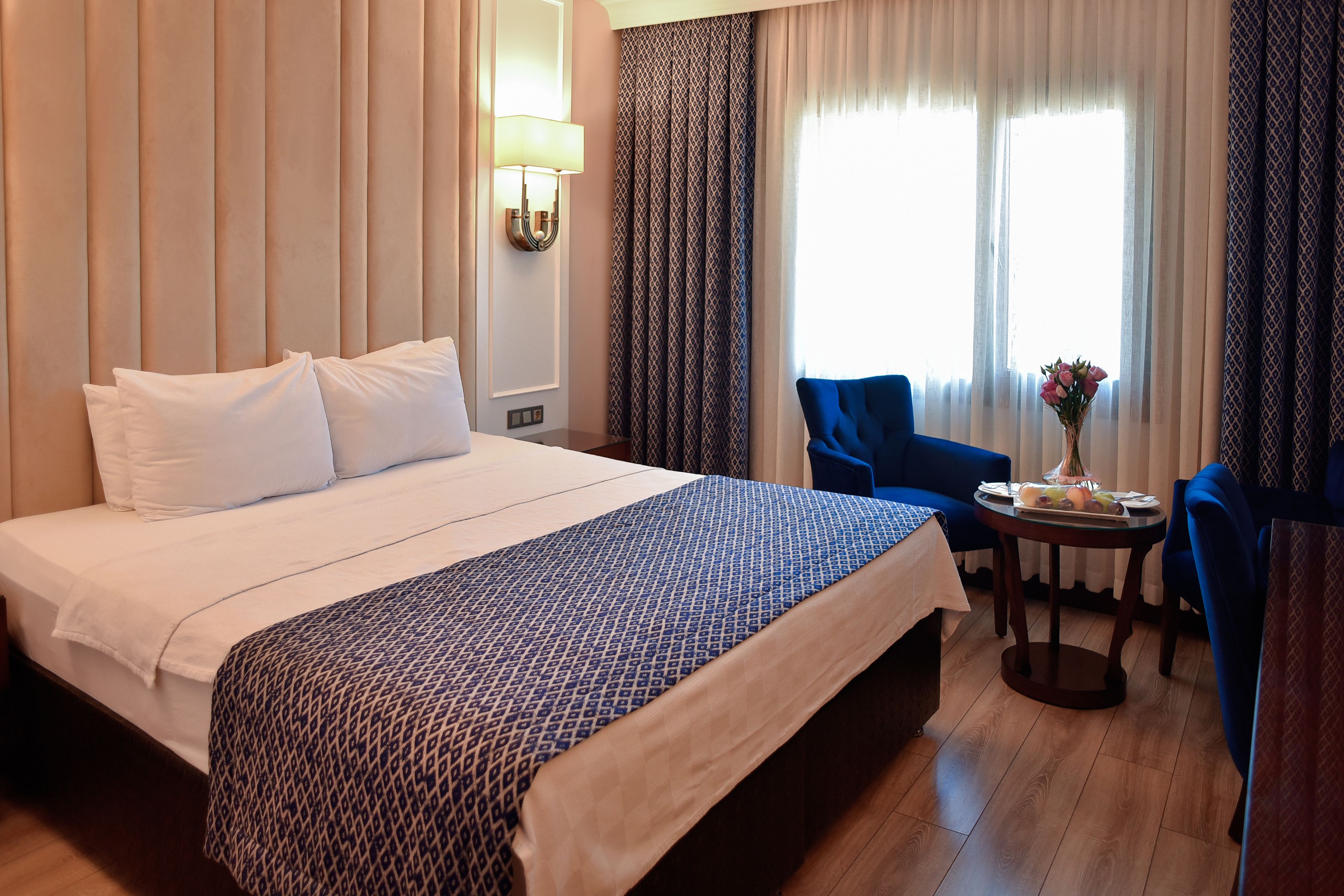 Habitación de hotel con una cama grande, una mesita con un jarrón de flores, un sillón azul y una ventana con cortinas.