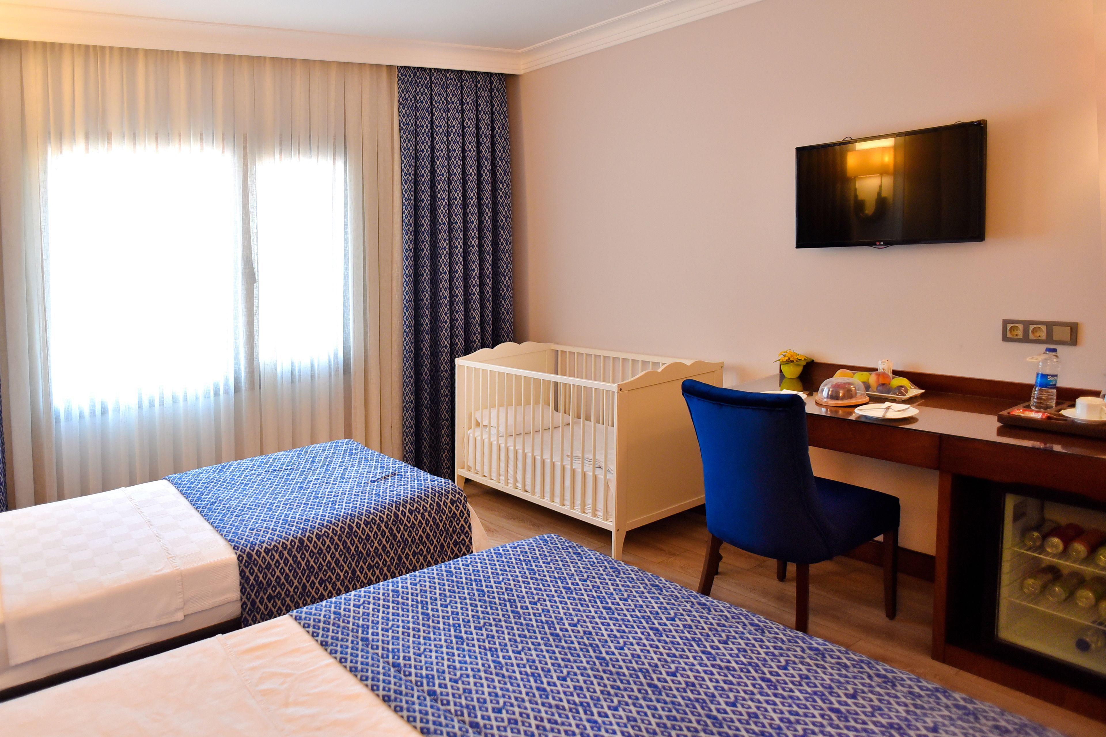 A hotel room with two beds, a crib, a desk with a chair, a TV, and a minibar.