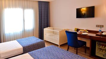 A hotel room with two beds, a crib, a desk with a chair, a TV, and a minibar.