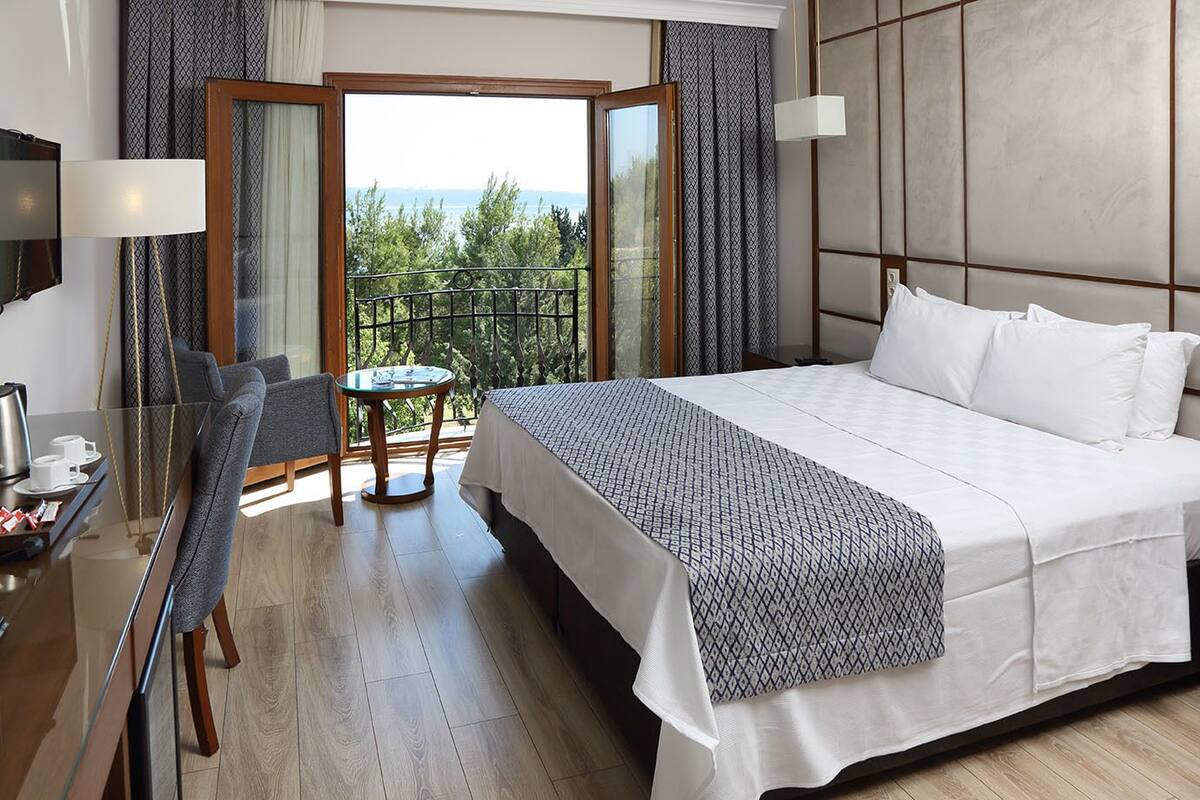 A hotel room with a large bed, a desk with a chair, a small table, and a balcony with a view of the outdoors.