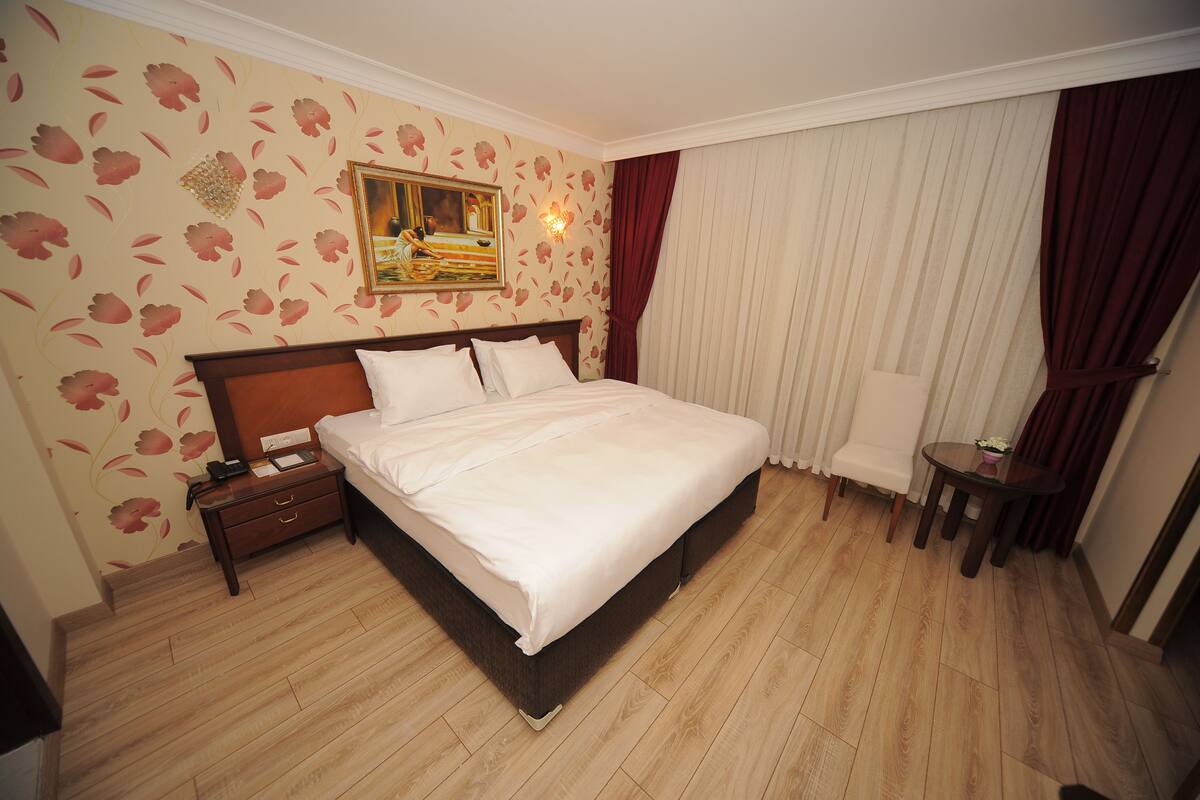 A hotel room with a large bed, a bedside table, a chair, and a small table with a vase.