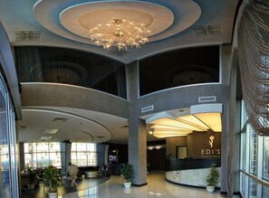 Un amplio vestíbulo de hotel con una lámpara de araña moderna, zonas de descanso y un mostrador de recepción.