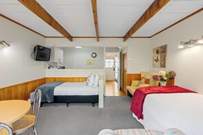 Habitación de hotel con techo de madera, una cama con una manta roja, una mesita con una silla, un televisor montado en la pared y un reloj en la pared.