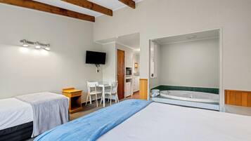 A hotel room with two beds, a desk, a TV, a kitchenette, and a bathroom with a bathtub.