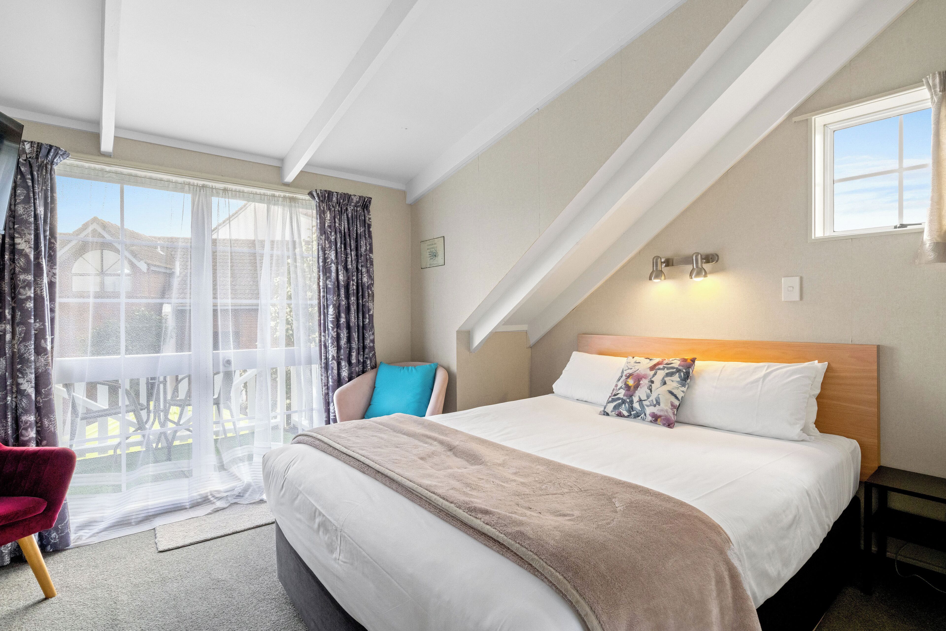A hotel room with a large bed, a chair, a window with curtains, and a view of a building outside.
