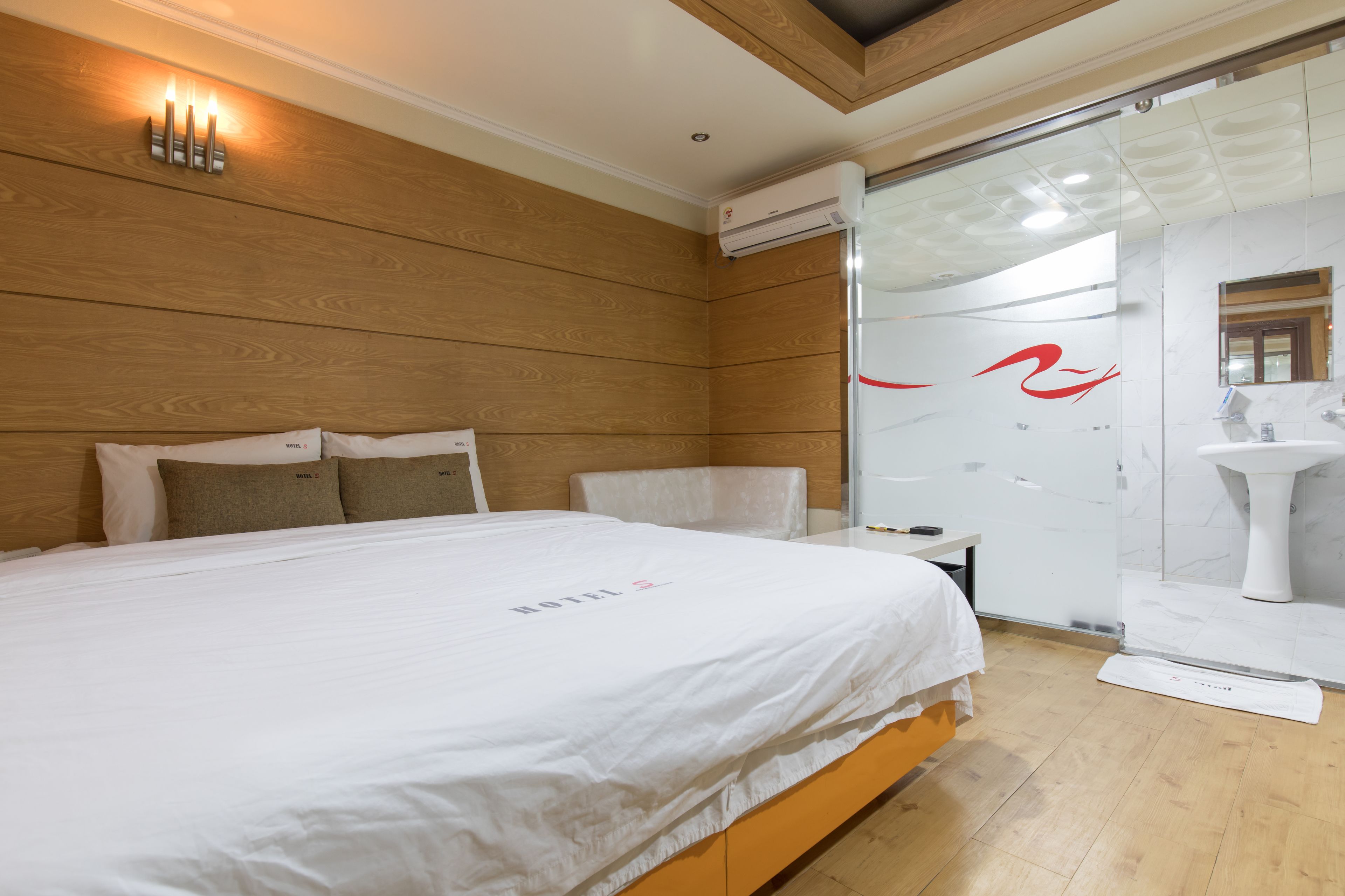 Une chambre d’hôtel avec une tête de lit en bois, un lit blanc recouvert d’une courtepointe blanche, une petite table de chevet et une cabine de douche en verre.