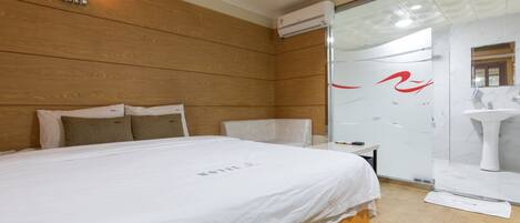 Une chambre dâhĂŽtel avec une tĂȘte de lit en bois, un lit blanc recouvert dâune courtepointe blanche, une petite table de chevet et une cabine de douche en verre.