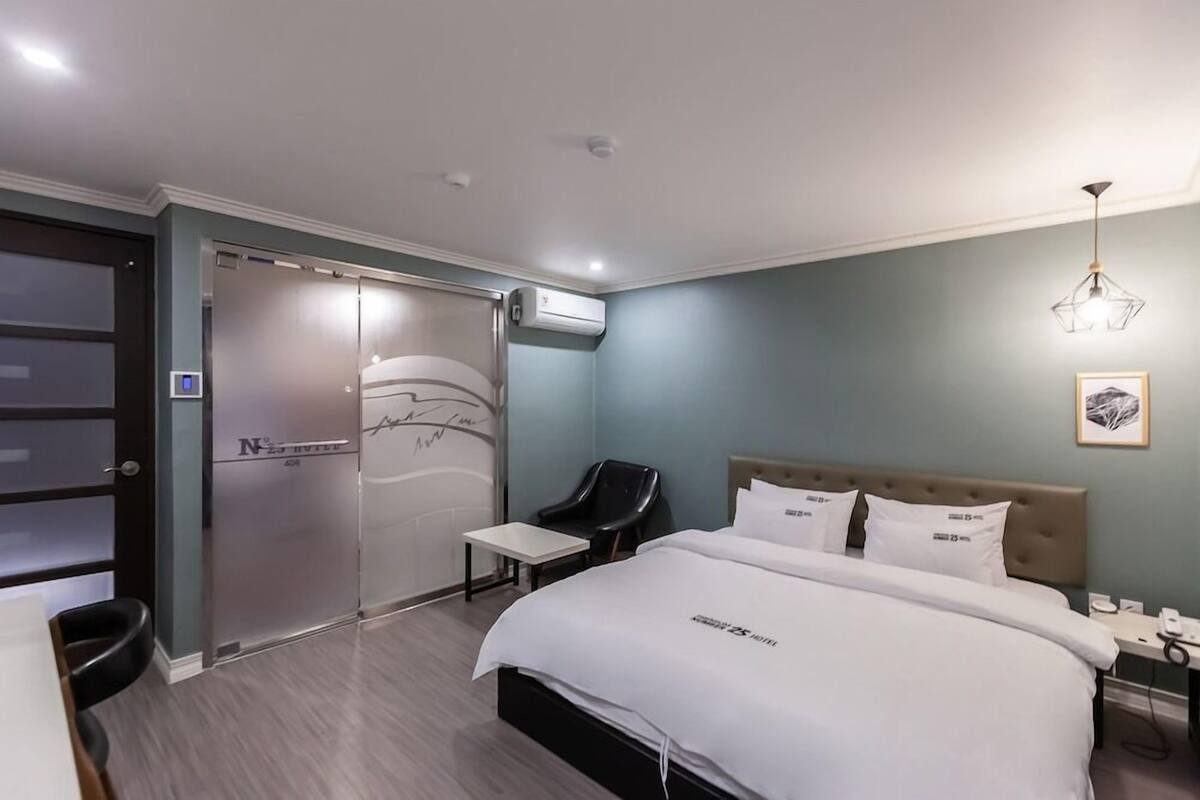 A hotel room with a bed, a chair, a desk, a wardrobe, and an air conditioning unit.