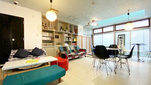A modern living room with a sofa, a small table, a bench, and a dining area with chairs and a table. There are shelves with books and decorative items, and a large window with blinds. - OUCHI HOTEL Yokogawa (Hiroshima)
