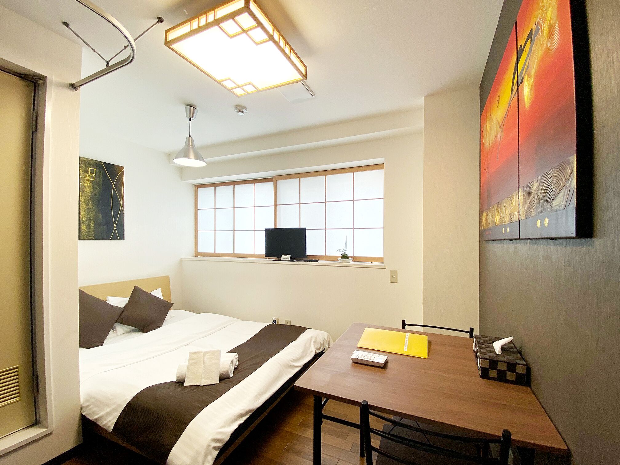 A hotel room with a bed, a desk, a window, a painting, and a ceiling light fixture.