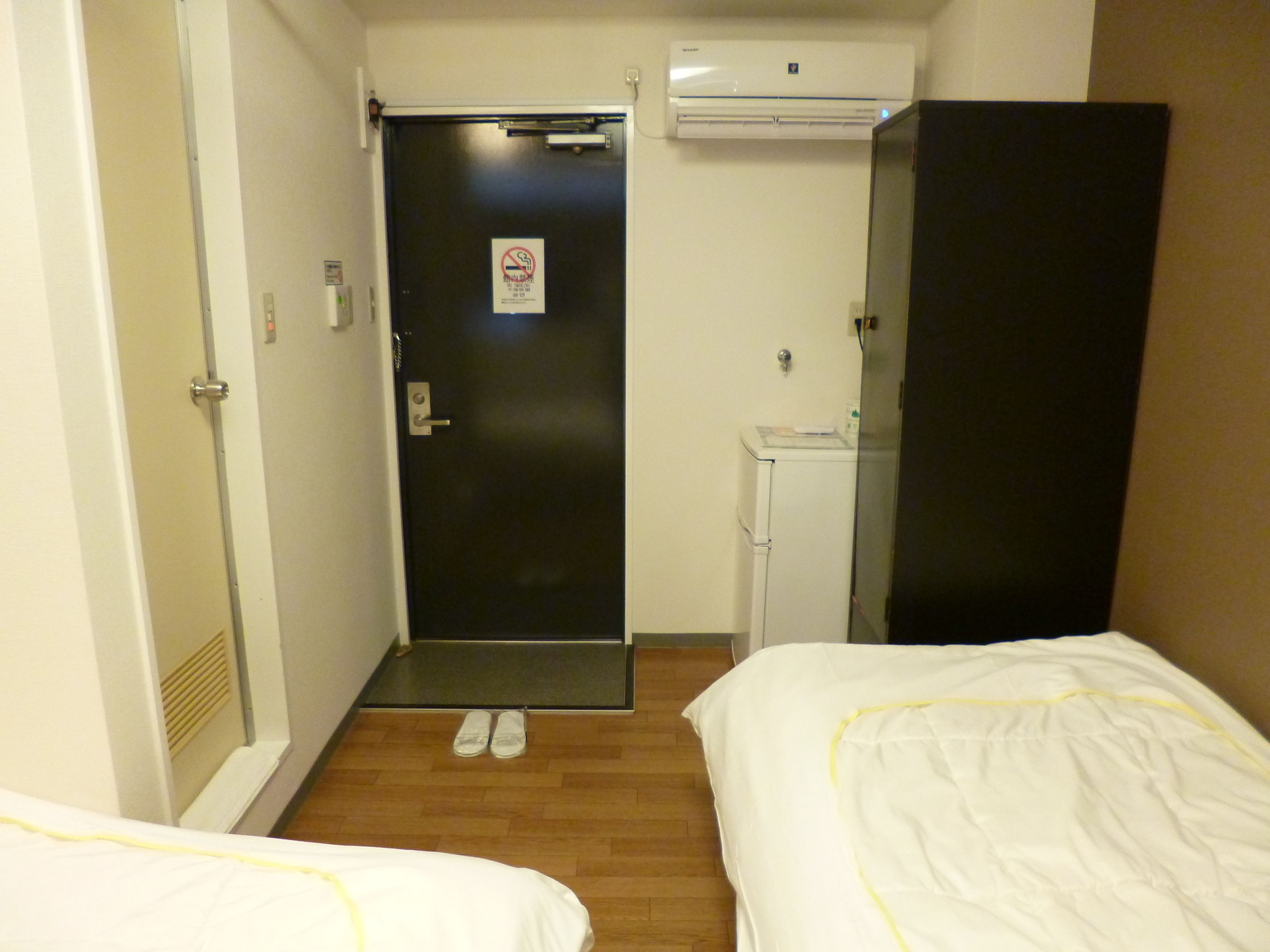 Habitación de hotel con dos camas, una puerta con cartel de no fumar, un equipo de aire acondicionado, un escritorio pequeño y un armario.