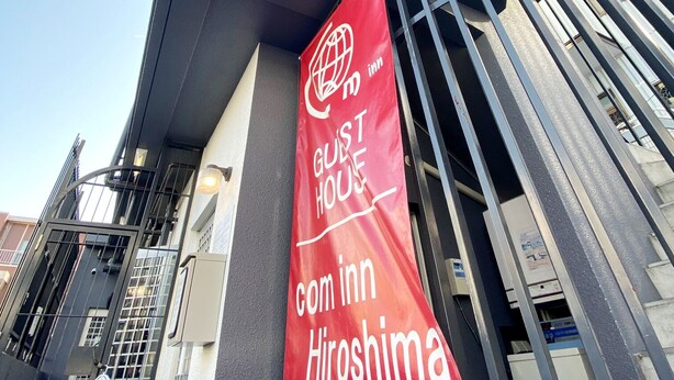 A modern building with a prominent red banner advertising a guest house named 'com inn Hiroshima'. - OUCHI HOTEL Yokogawa (Hiroshima)