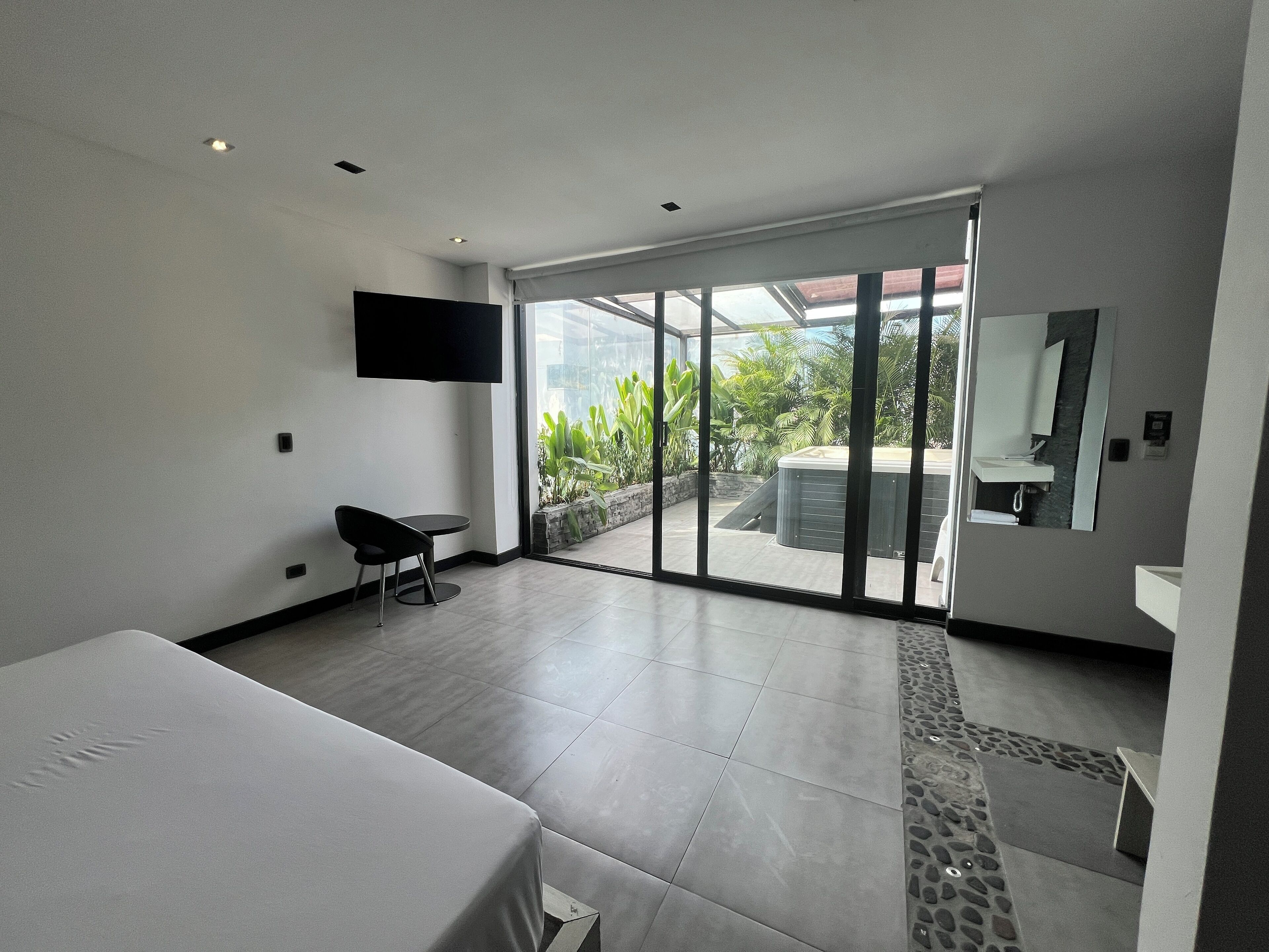 Habitación de hotel moderna con un amplio ventanal corredero que da a un balcón con vistas a zonas verdes. Se aprecia un televisor de pantalla plana en la pared, una pequeña mesa con una silla y una cama con cubrecama blanco.