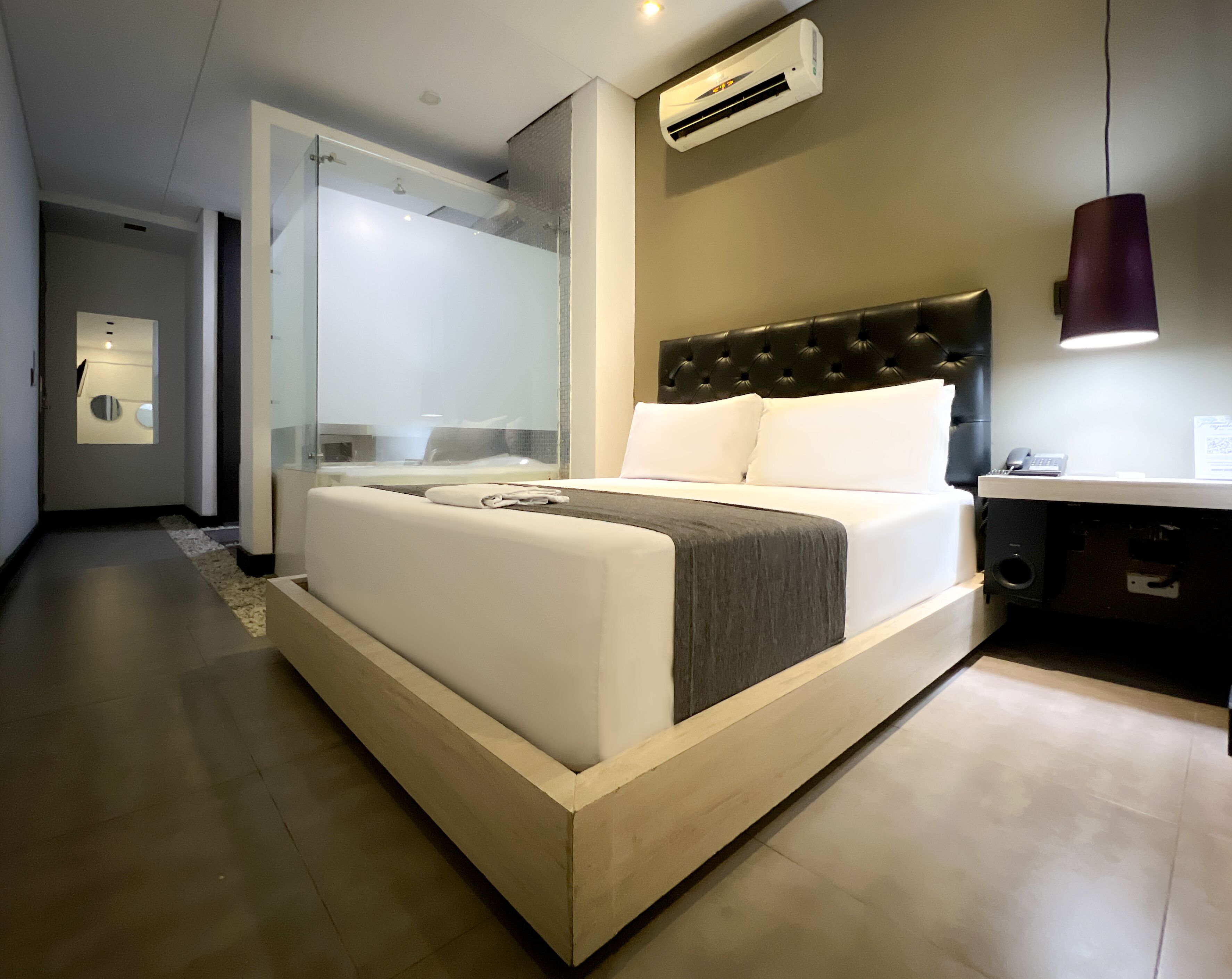 A modern hotel room with a large bed, a bedside table, a desk, and a bathroom visible through a glass partition.