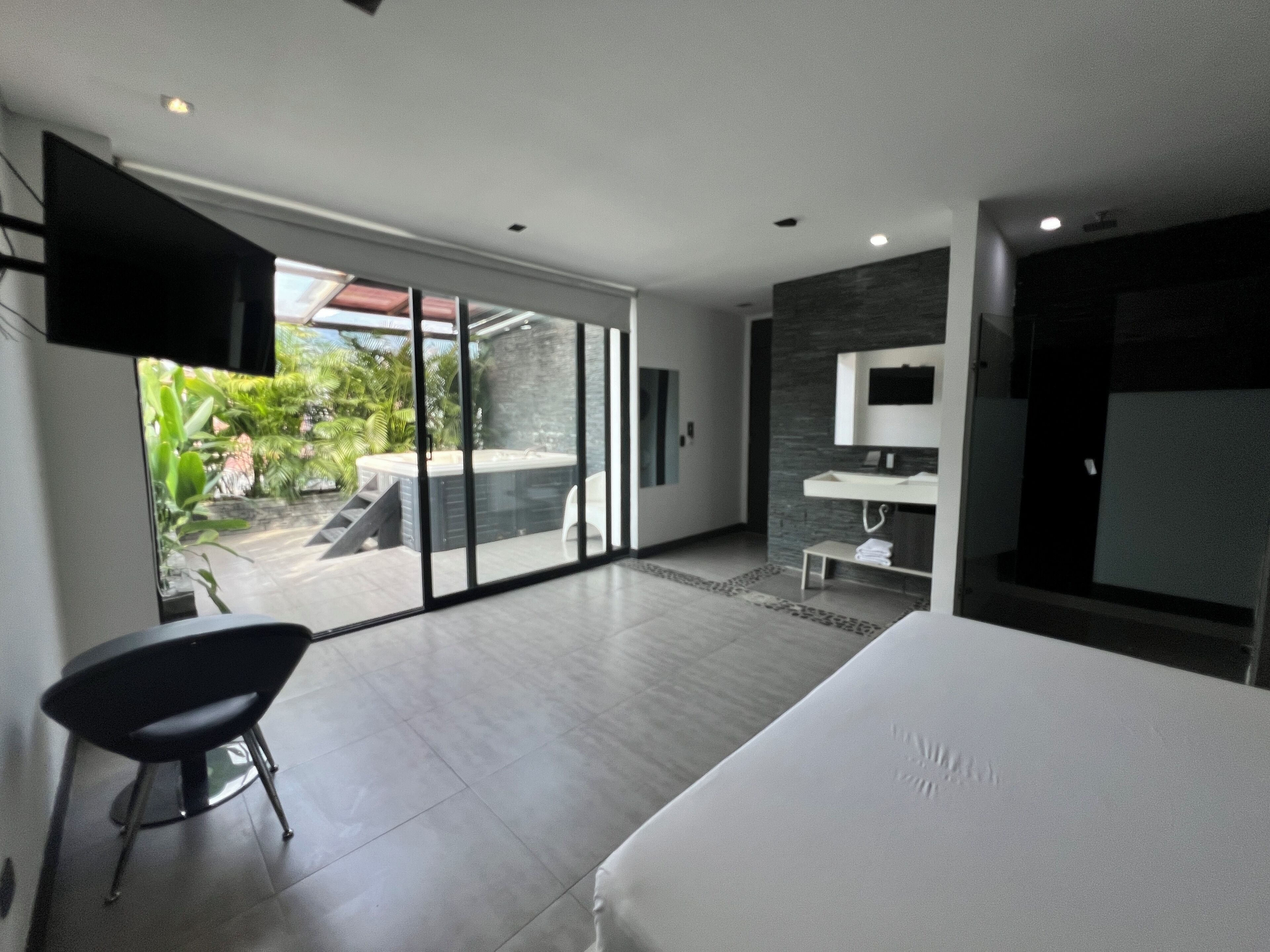 Un dormitorio moderno con una cama grande, un televisor montado en la pared y un ventanal corredero que da a un balcón con jacuzzi.