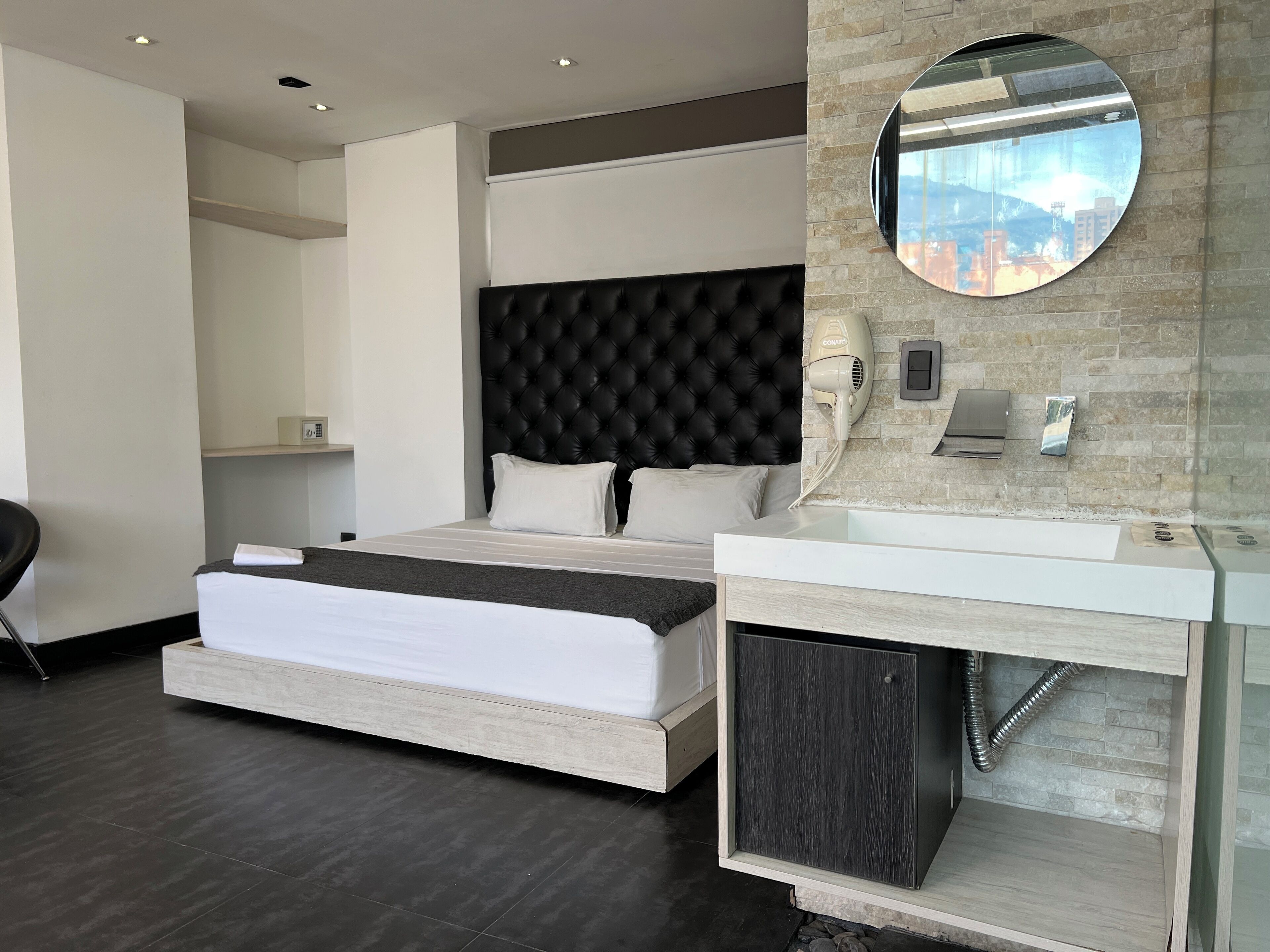A modern hotel room with a large bed, a round mirror, a built-in vanity, and a city view.