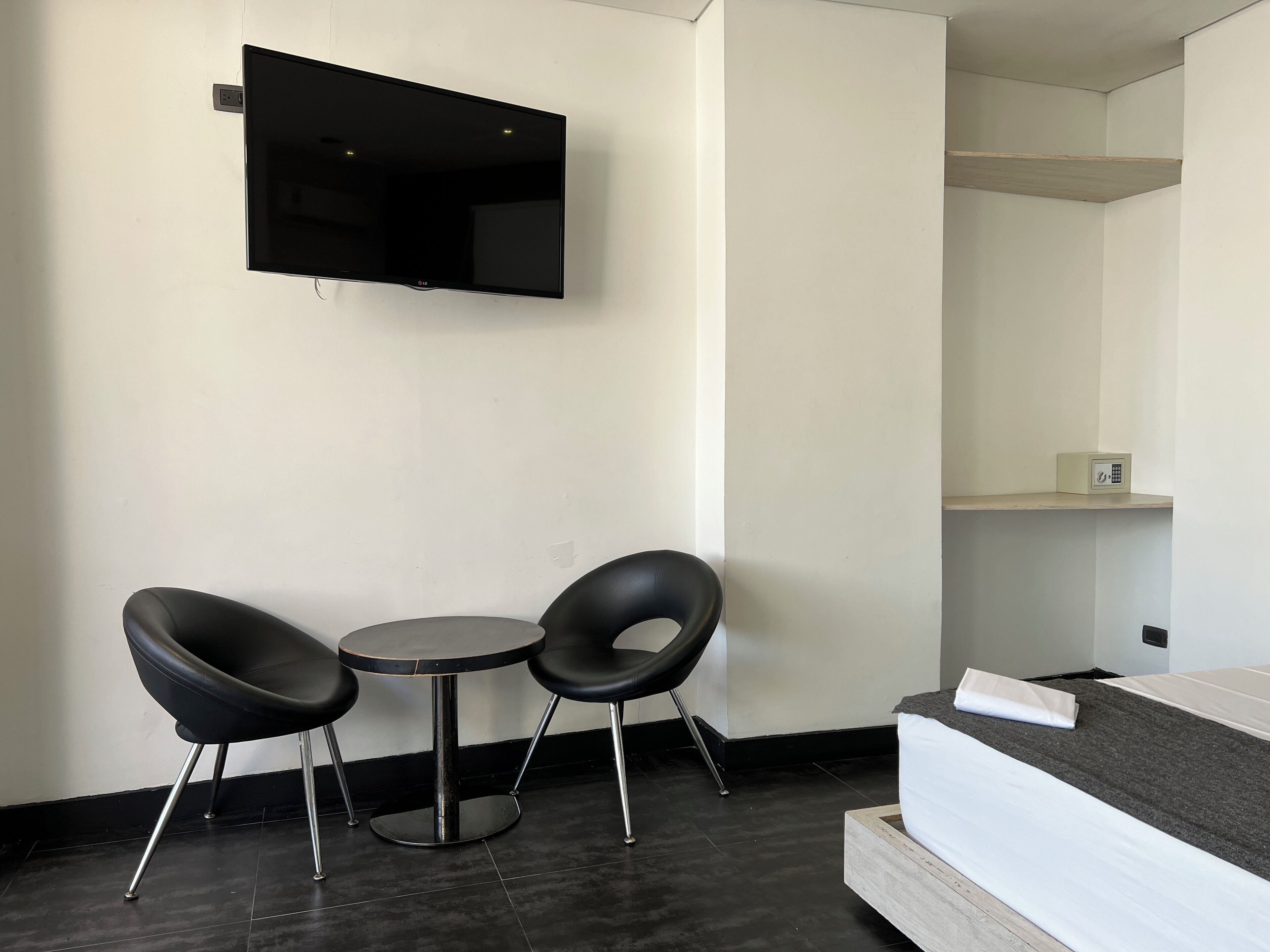 Una habitación moderna con televisión de pantalla plana en la pared, una pequeña mesa redonda con dos sillas negras, y una cama con colchón gris y sábanas blancas.