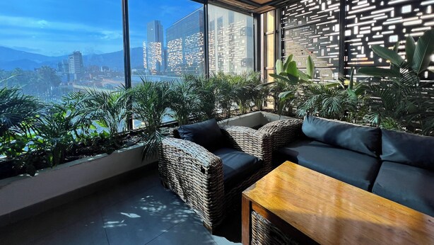 A modern interior with wicker furniture, a wooden table, and large windows offering a city view. - Hotel 47 Medellin Street (Medellín)