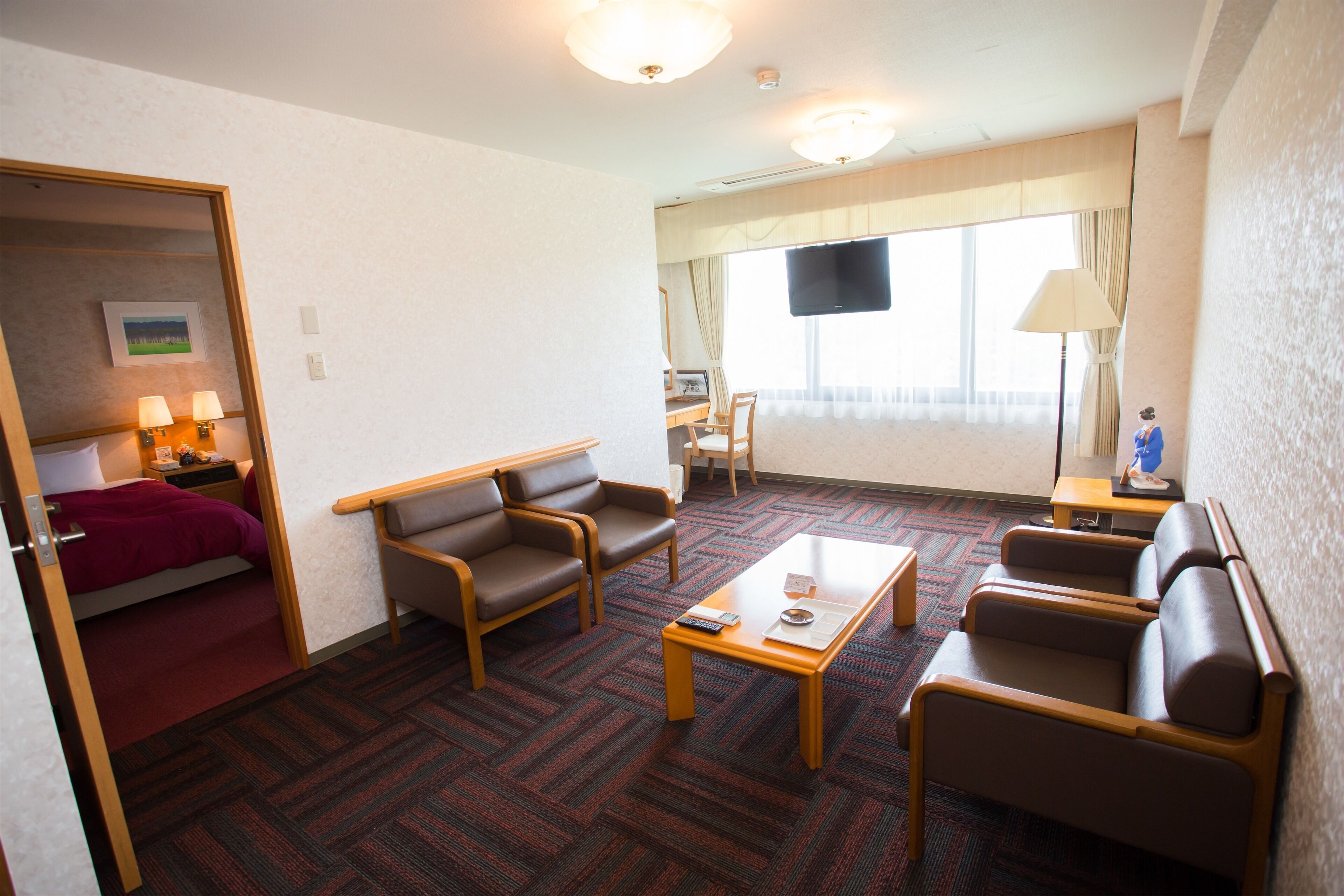 Ein Hotelzimmer mit einem Bett, zwei Sesseln, einem Sofa, einem Couchtisch, einem Schreibtisch mit Stuhl, einem Fernseher und einem Fenster mit Vorhängen.