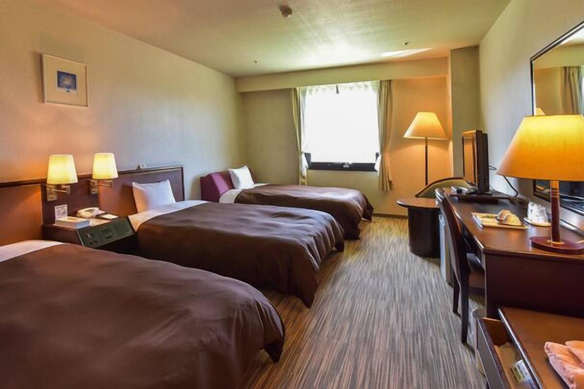 Quarto de hotel com duas camas, uma escrivaninha, uma cadeira, um abajur e uma janela.