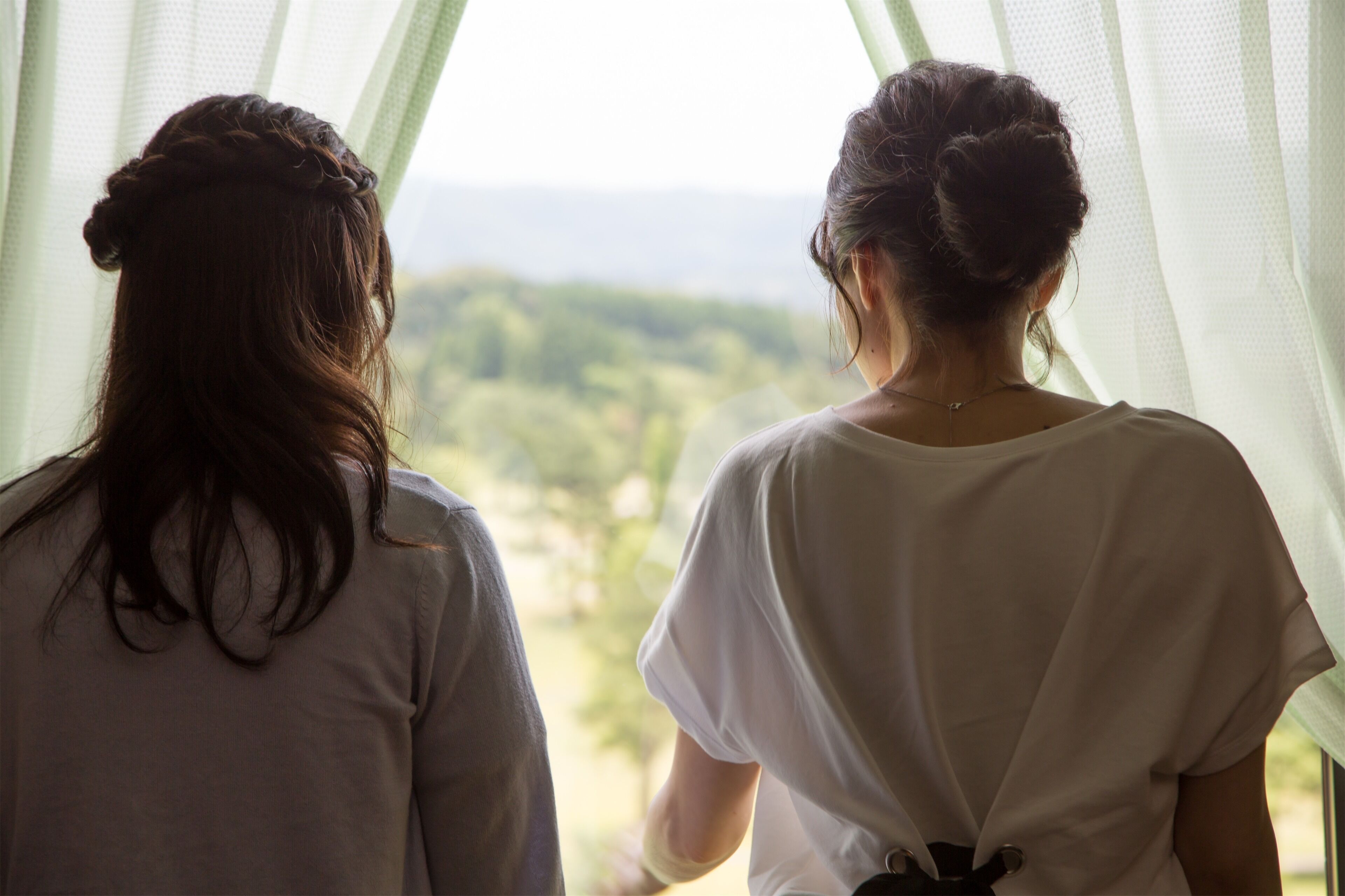 Zwei Personen stehen mit dem Rücken zur Kamera und blicken aus einem Fenster auf eine grüne Landschaft.