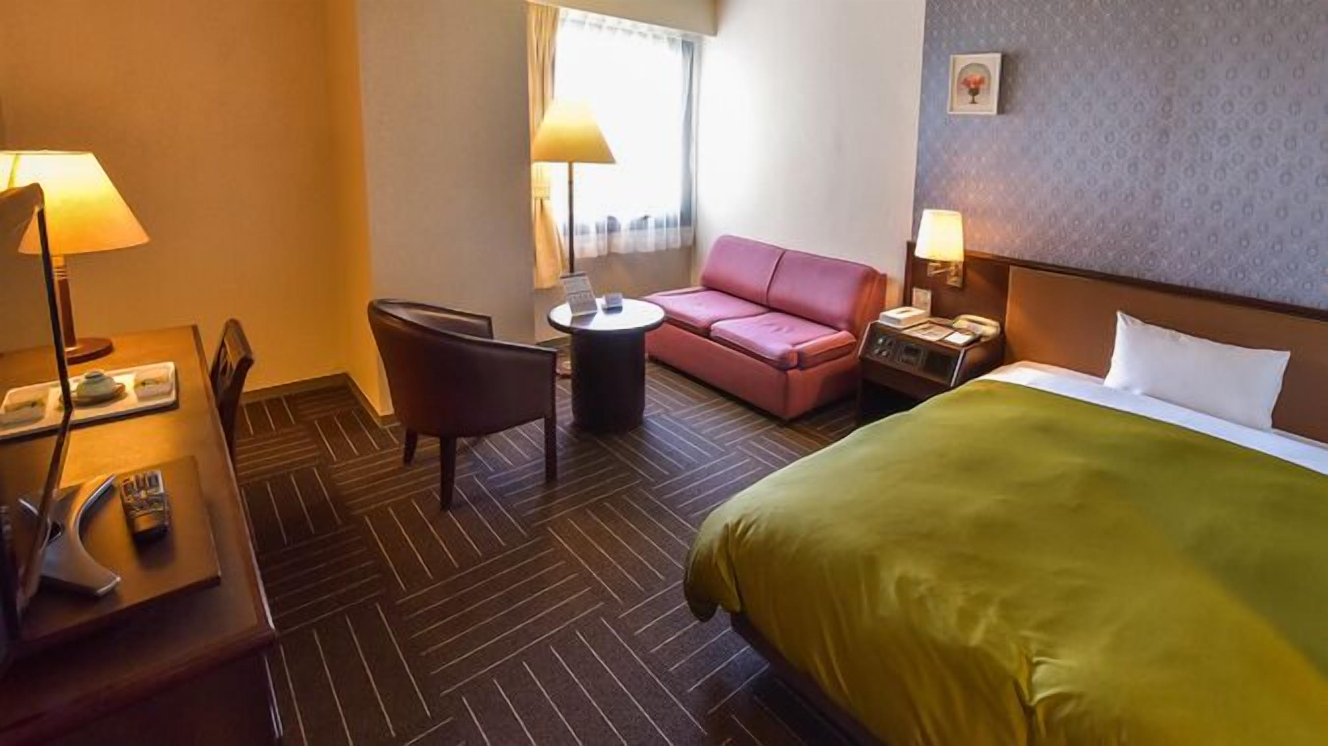 A hotel room with a bed, a desk with a lamp, a chair, a small table, and a pink sofa.